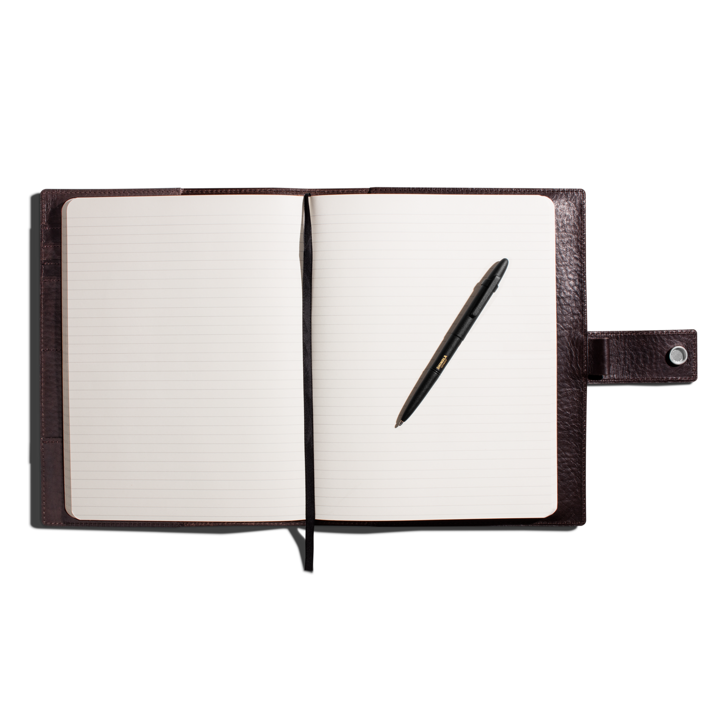 A leather-bound notebook with lined pages, a Shinola + Fisher Bullet Space Pen - Matte Black from Shinola resting on the right page, and a buttoned strap closure on the right, all set against a white background.