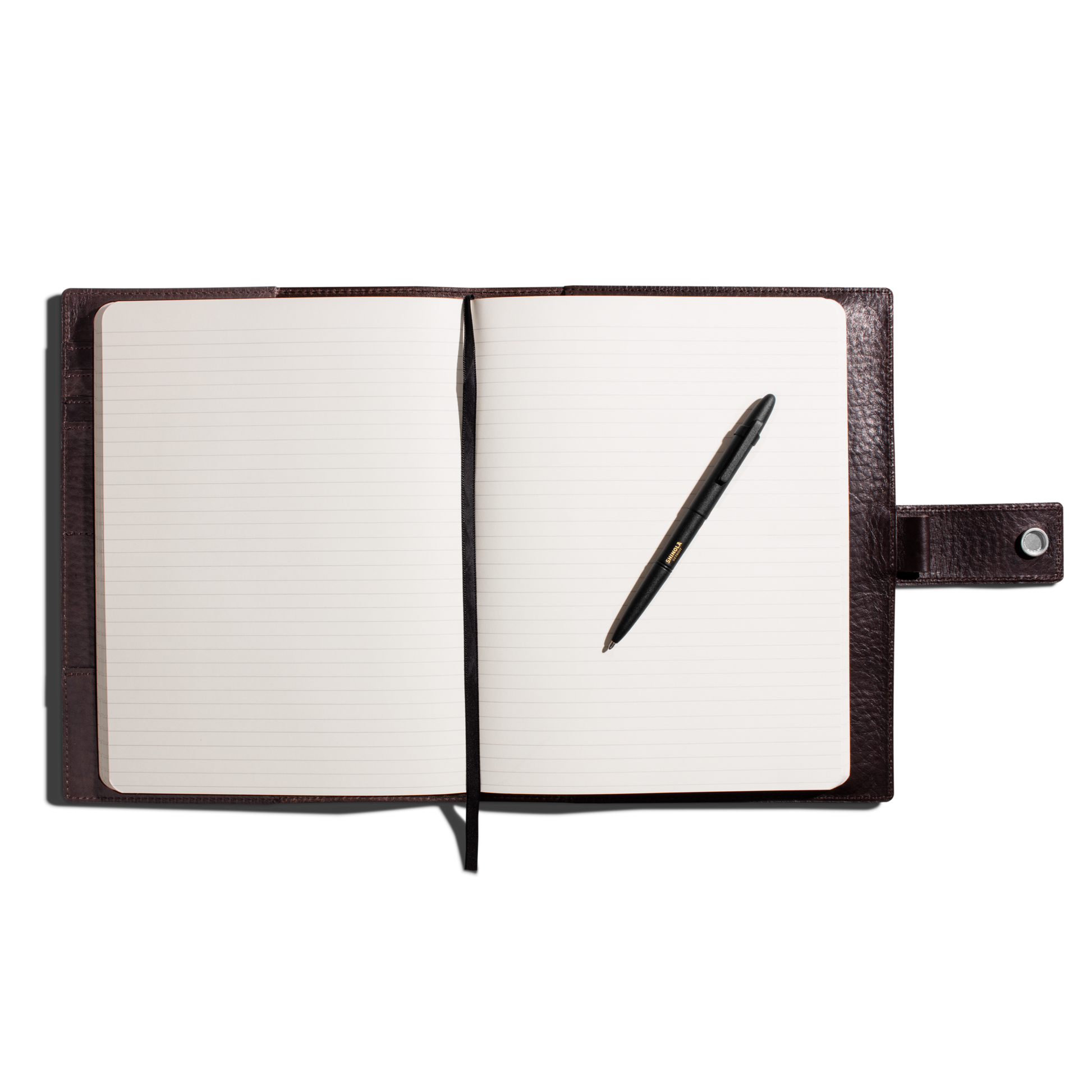 A leather-bound notebook with lined pages, a Shinola + Fisher Bullet Space Pen - Matte Black from Shinola resting on the right page, and a buttoned strap closure on the right, all set against a white background.