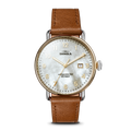 The Shinola Canfield Watch features a tan leather strap, round gold-tone case, white mother-of-pearl dial with gold numerals, and a date window at 3 o’clock. The face displays “Shinola” and “Argonite-705, Detroit.”.