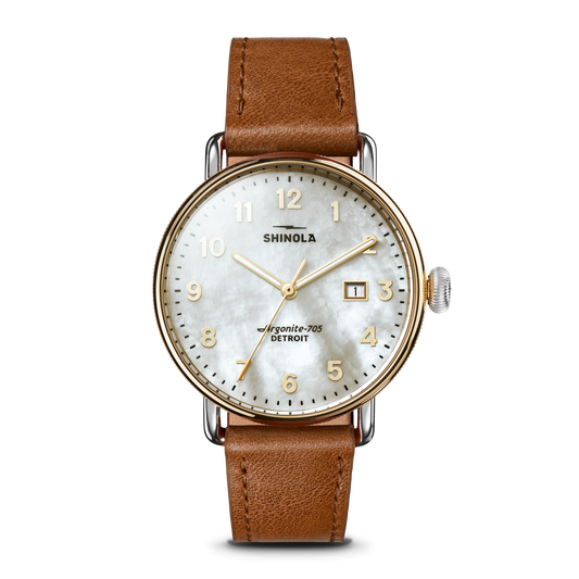 The Shinola Canfield Watch features a tan leather strap, round gold-tone case, white mother-of-pearl dial with gold numerals, and a date window at 3 o’clock. The face displays “Shinola” and “Argonite-705, Detroit.”.