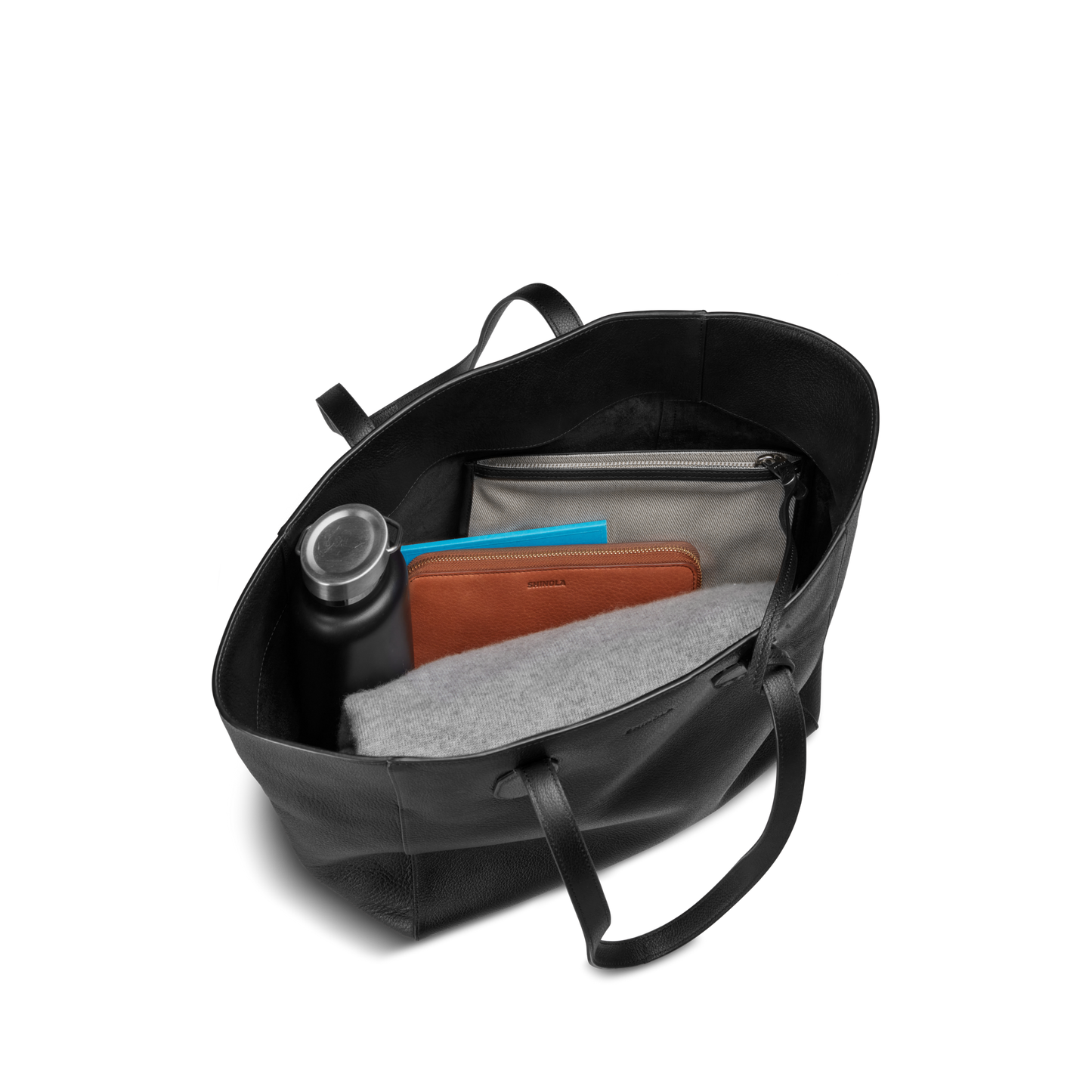 The Shinola Runabout Tote Bag - Black is a lightweight pebbled leather tote with two handles, holding a black water bottle, brown zipper pouch, gray felt item, and gray zippered pouch containing a blue notebook.