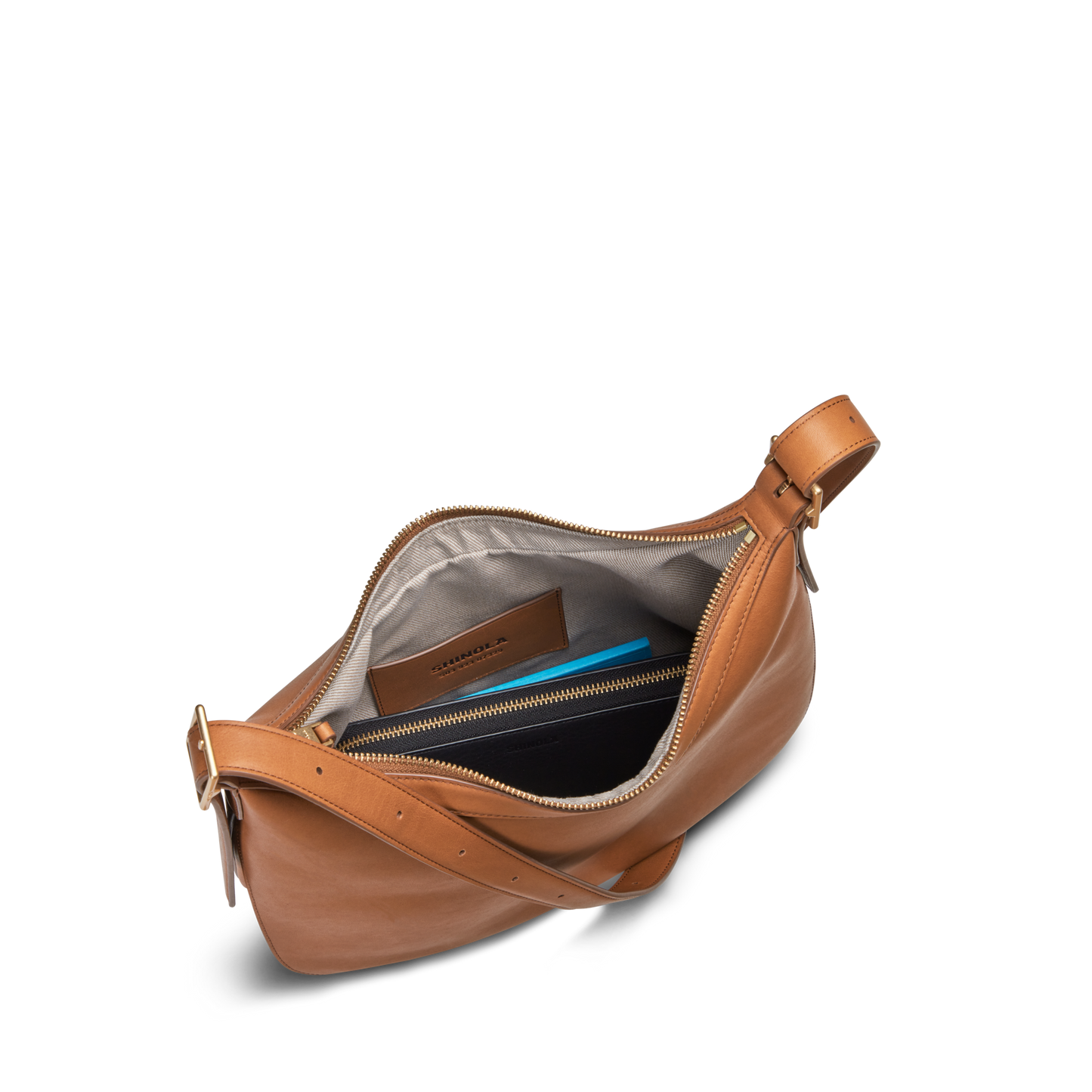 The Shinola Bixby Shoulder Bag - Tan features natural leather, a top zipper, gray lining, two inner pockets for a brown wallet and blue notebook, plus an adjustable buckle strap and accordion-style design for style and function.