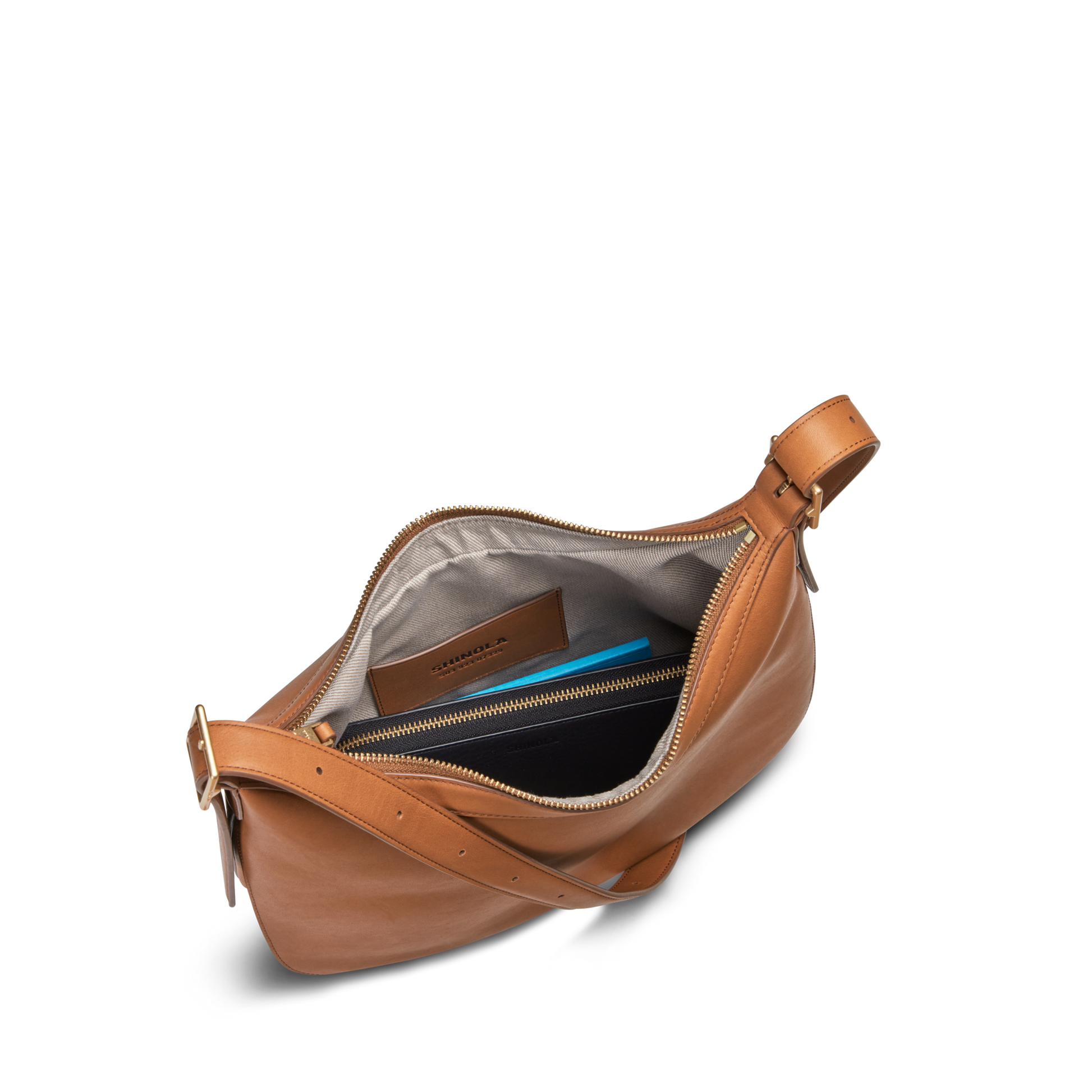 The Shinola Bixby Shoulder Bag - Tan features natural leather, a top zipper, gray lining, two inner pockets for a brown wallet and blue notebook, plus an adjustable buckle strap and accordion-style design for style and function.