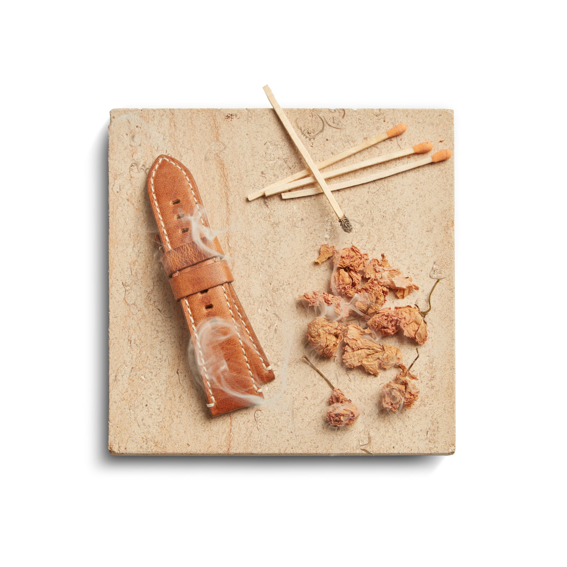 A tan leather watch strap, dried flower petals, and burnt matches rest on a beige stone tile beside a Shinola Hotel 8oz Scented Candle in a recycled glass vessel. White wisps, possibly feathers or smoke, are scattered among the objects.