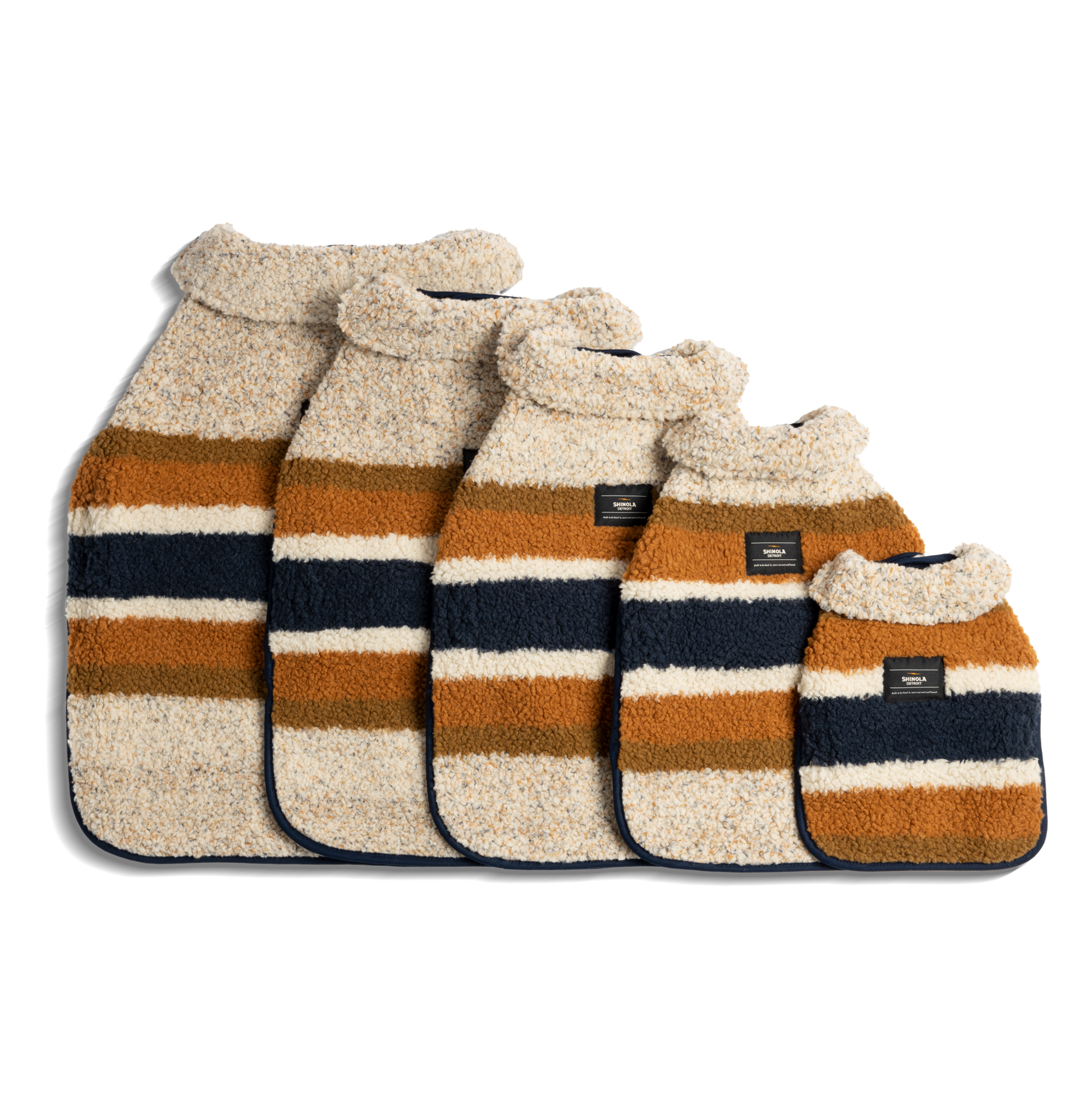 Five Shinola "Dog Coat - Brand Stripe Ombre" hot water bottles, each in a different size, feature fluffy striped covers in beige, brown, and navy blue and are arranged from largest to smallest on a white background.