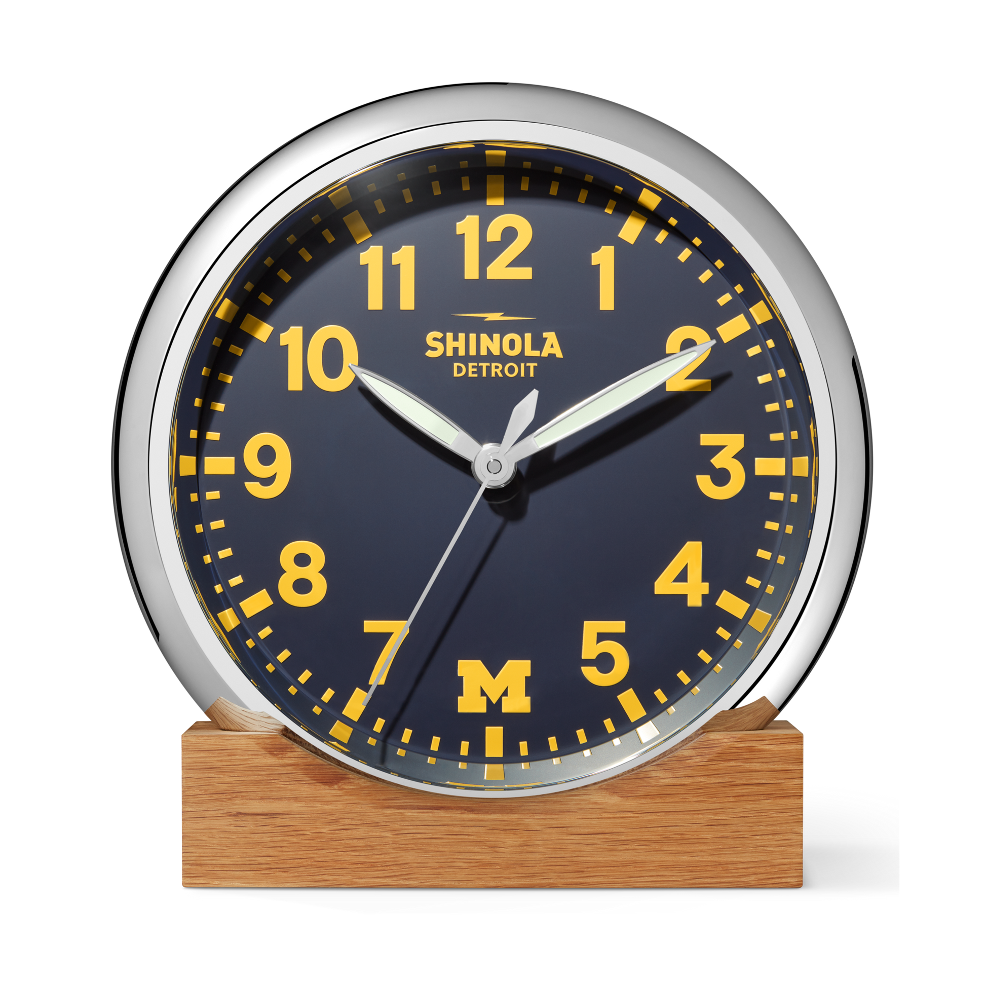 The 14" Runwell Clock - Blue by Shinola features a round analog design with a navy face, yellow numbers and hands, and "Shinola Detroit" branding. A yellow M on the face represents the University of Michigan. The clock sits on a wooden base.