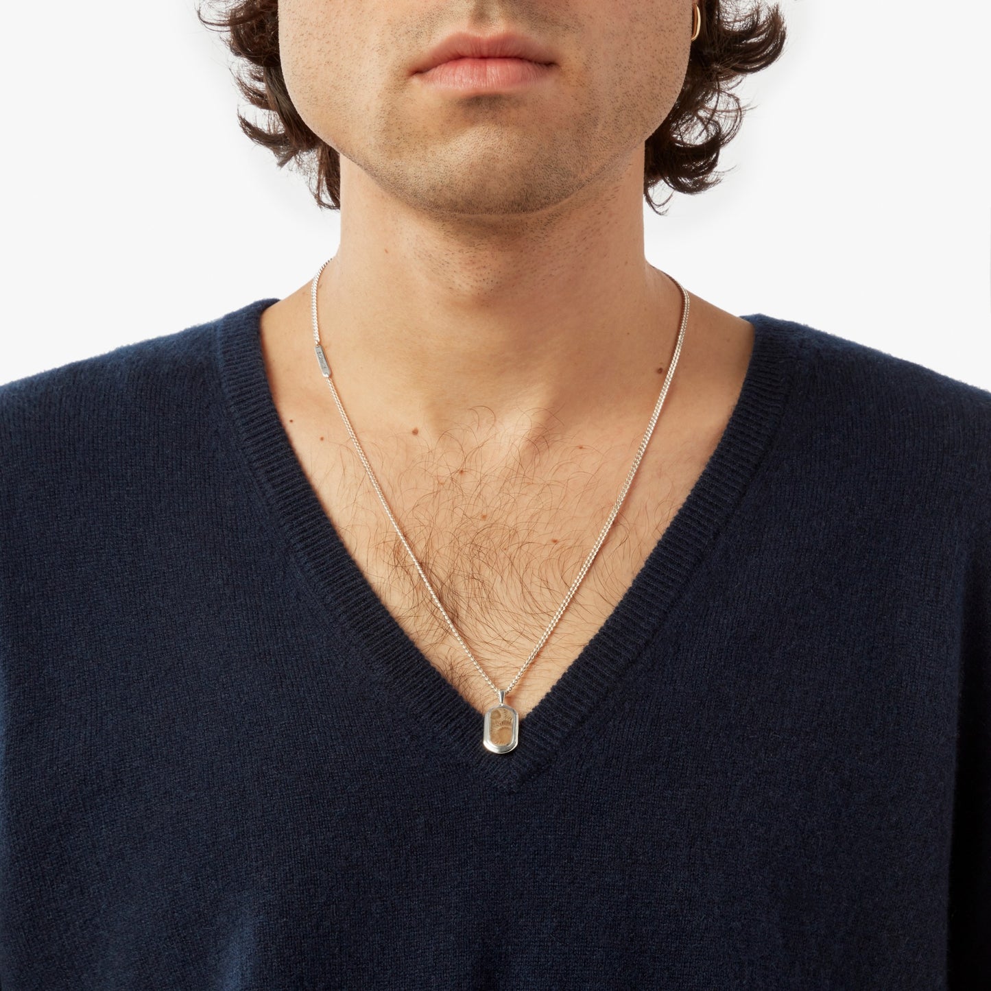 A person wears a dark blue V-neck sweater and a Shinola Petoskey Tag Necklace in sterling silver, shown from shoulders to just above the lips against a plain white background.
