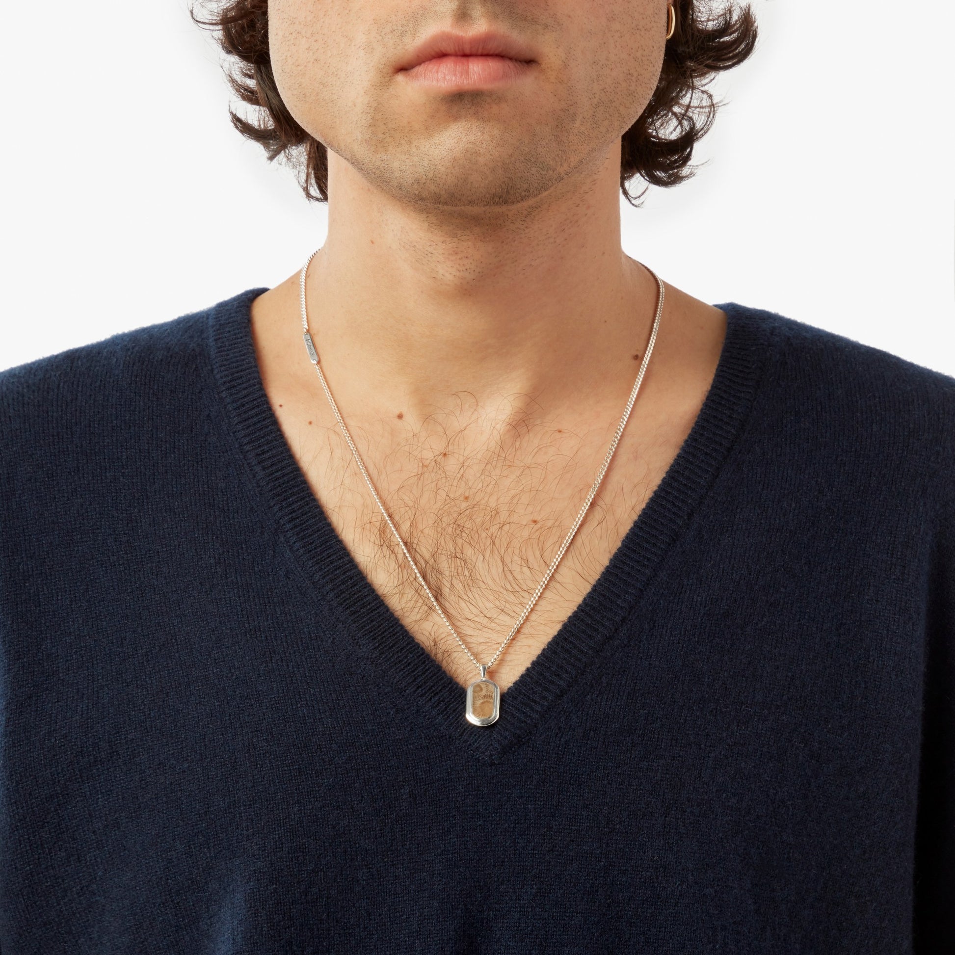 A person wears a dark blue V-neck sweater and a Shinola Petoskey Tag Necklace in sterling silver, shown from shoulders to just above the lips against a plain white background.