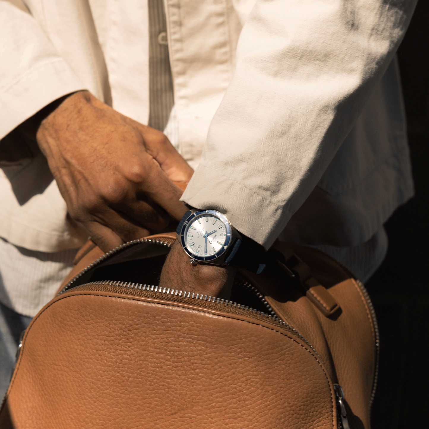 Wearing a white shirt and the Shinola Duck Watch - Midnight Blue, someone reaches into a brown leather bag as sunlight casts shadows across their hand and clothing.