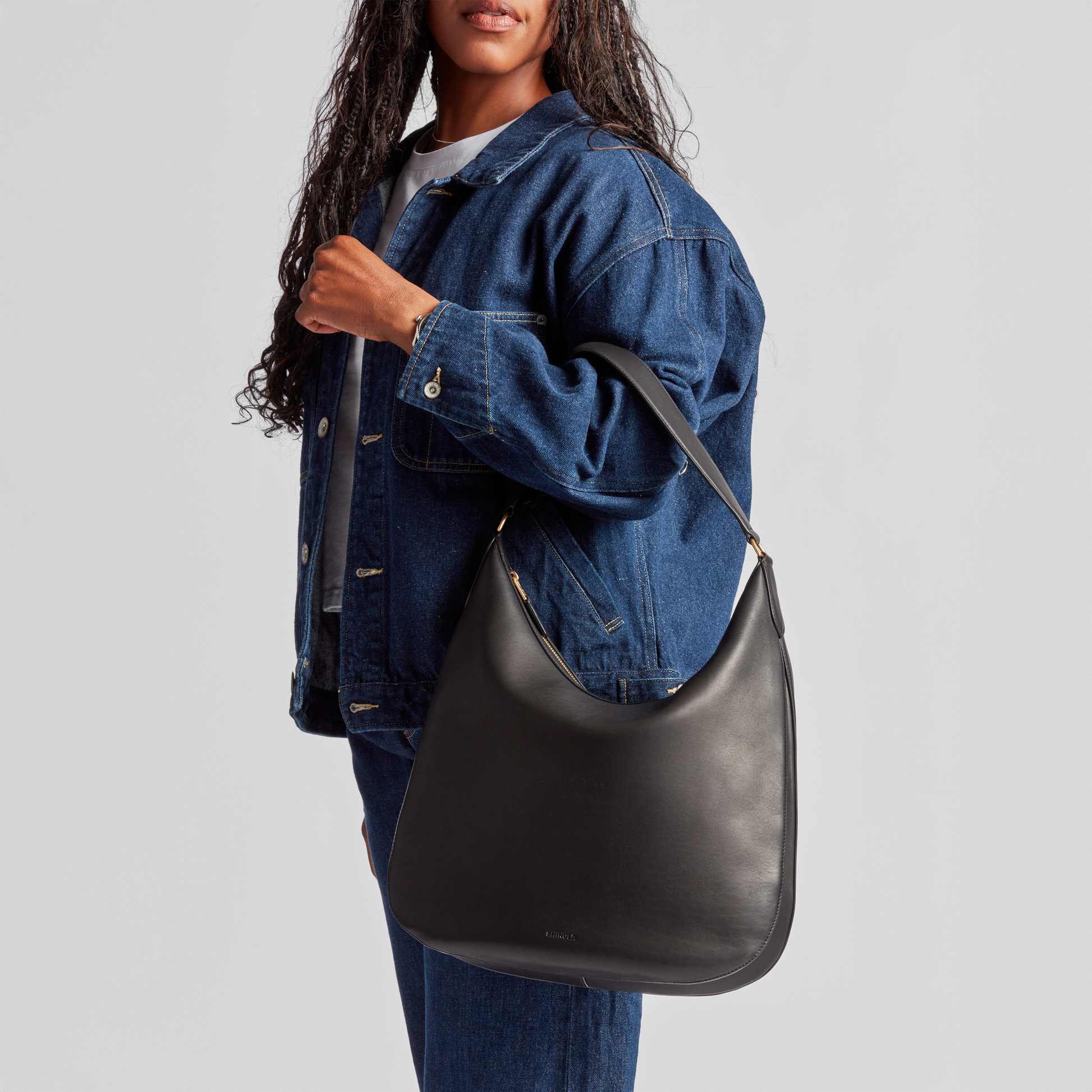 A woman in a blue denim jacket and jeans holds the Shinola Bixby Shoulder Bag - Black, featuring natural leather and accordion-style construction, hanging on her shoulder against a plain light gray background.