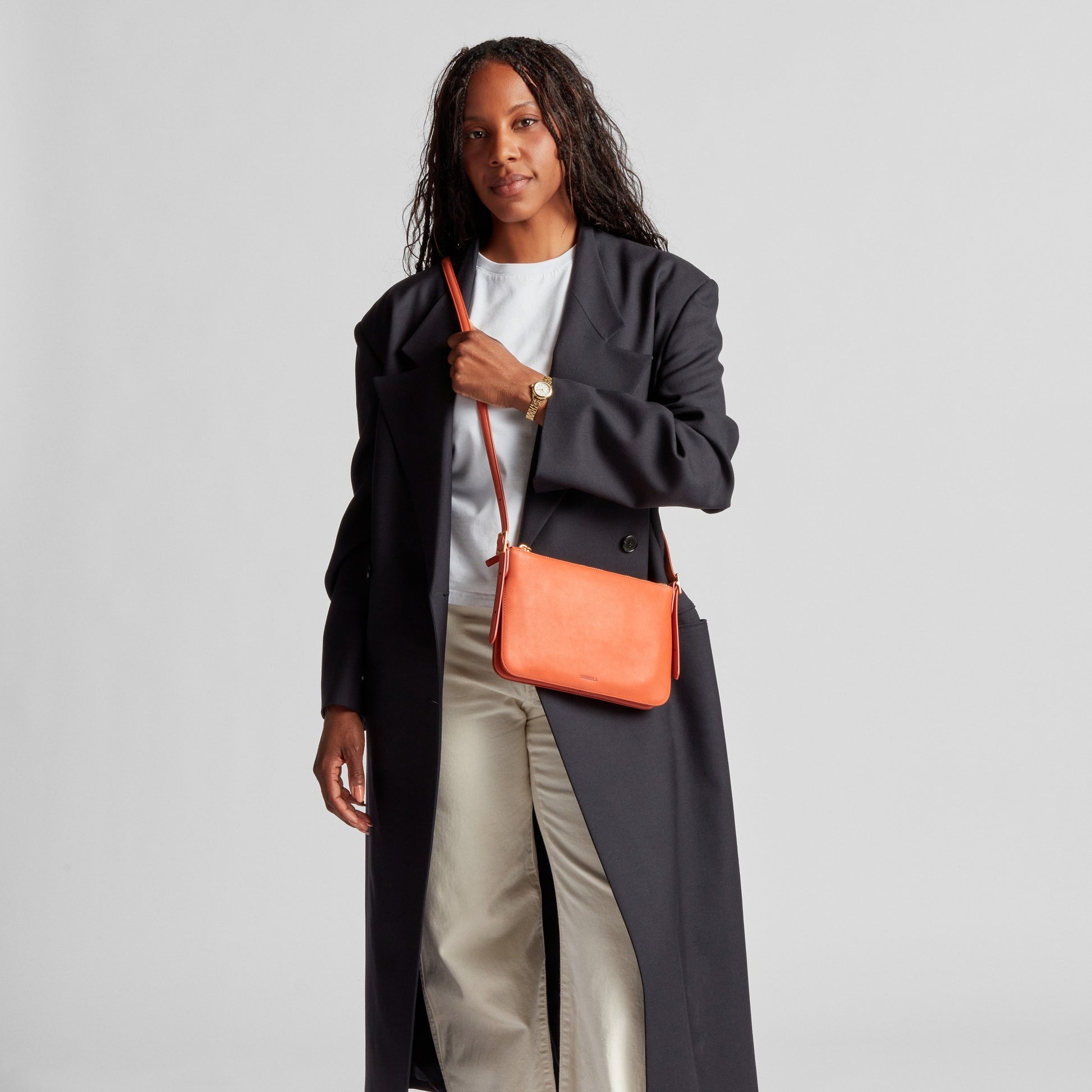 A woman in a long dark coat and light pants stands against a plain background, holding the Shinola Small Bixby Crossbody in Rust—an everyday accessory—and looks at the camera.