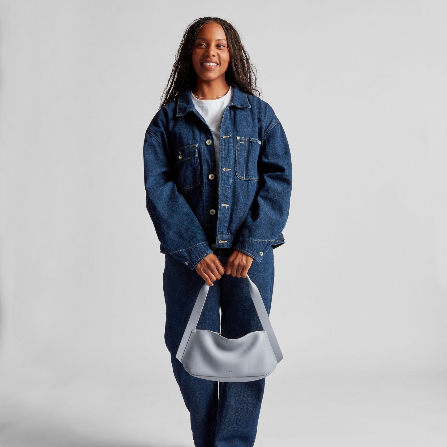 A person in a blue denim jacket and jeans stands against a plain light background, smiling and holding the Shinola Small Derby Shoulder Bag - Mist with both hands in front of them.