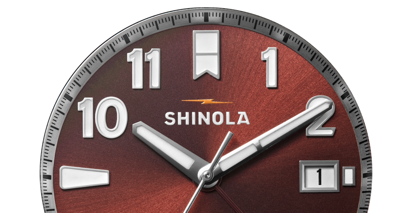 Close-up of the Shinola Monster GMT Automatic Watch - Whiskey, featuring a brown metallic face, silver hands, bold white numerals, a date window showing "1," and a yellow lightning bolt—an ideal travel companion with automatic movement.