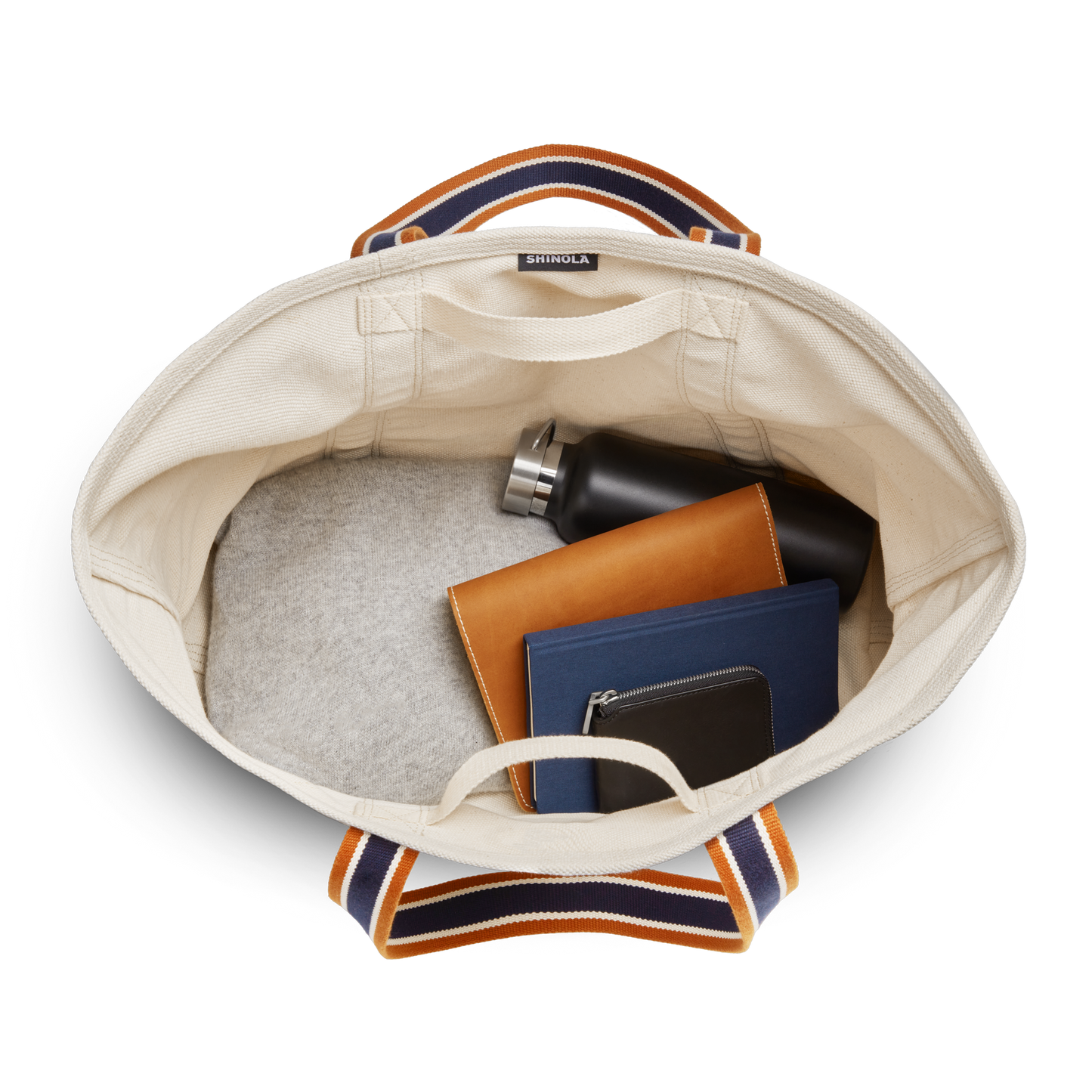 Top view of the Shinola Large Easy Canvas Tote - Natural with striped handles, showing a spacious open compartment and exterior slip pocket. Inside: black water bottle, brown notebook, blue book, and black zippered pouch.
