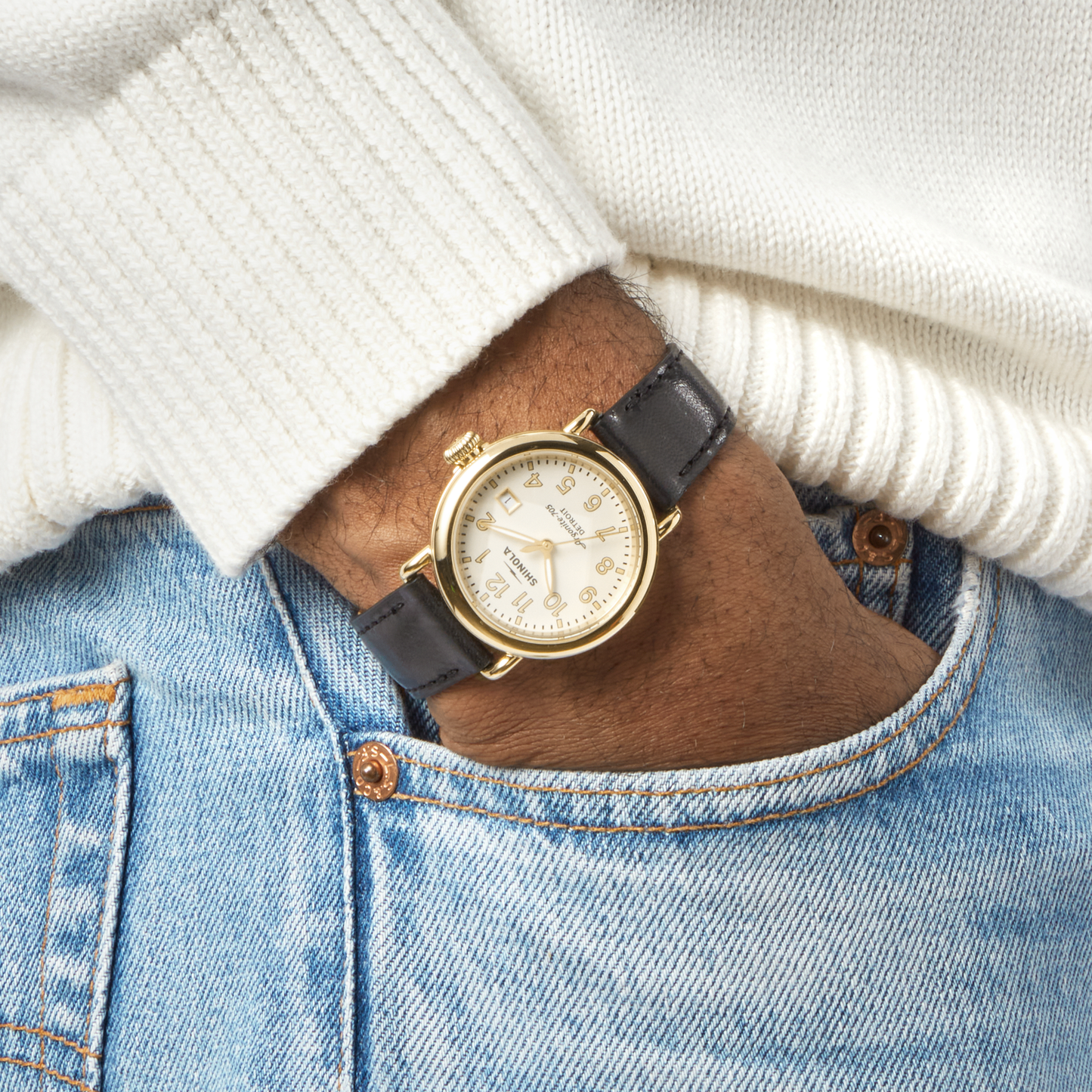 A person in a white sweater and blue jeans has their hand in their pocket, showcasing the Shinola Runwell Watch - Alabaster with a gold stainless steel case and black leather strap on their wrist.