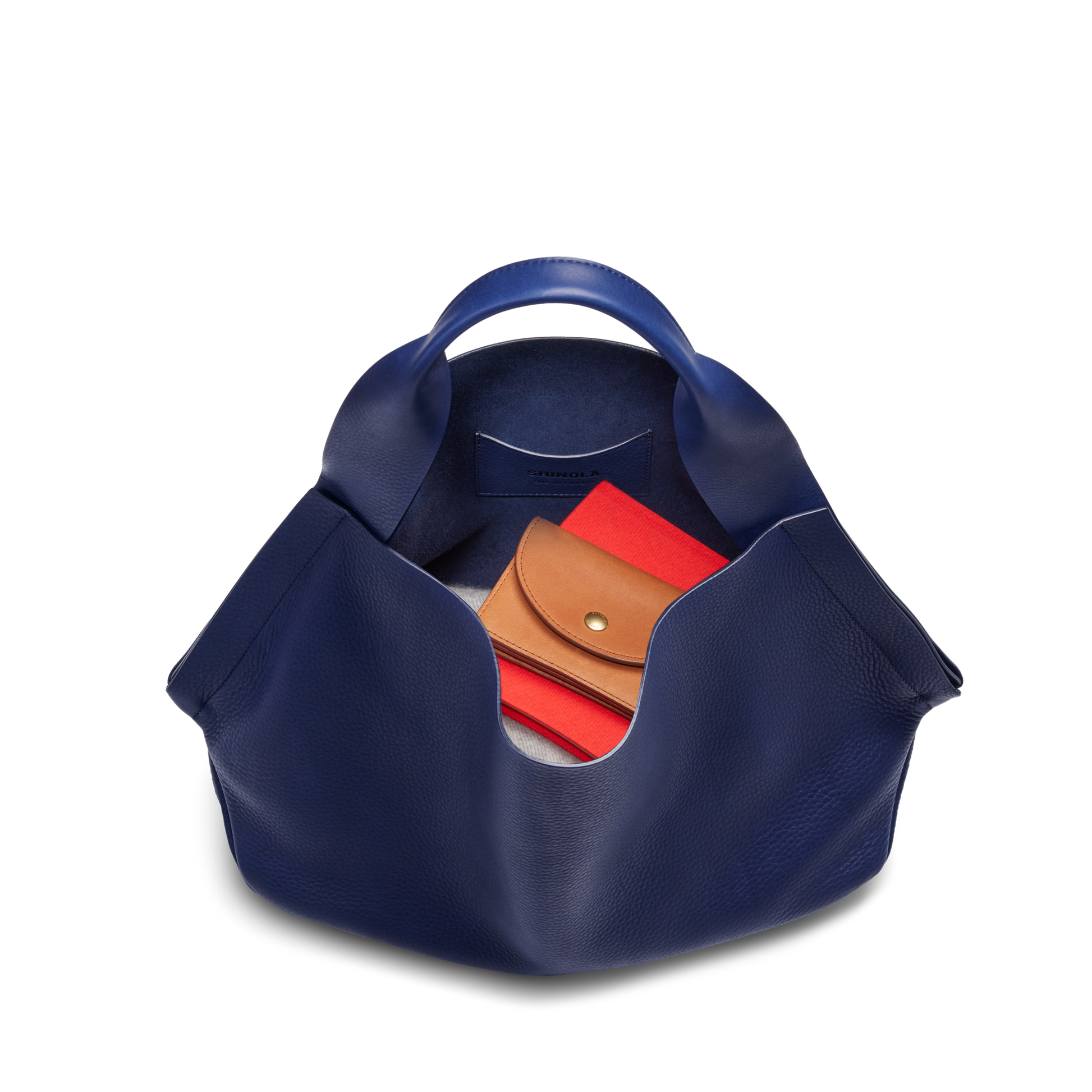 A top view of the Shinola Runwell Shoulder Bag - Lake shows its wide opening with a tan wallet in the leather card pocket and a red item inside, plus sturdy handles—all set against a white background.
