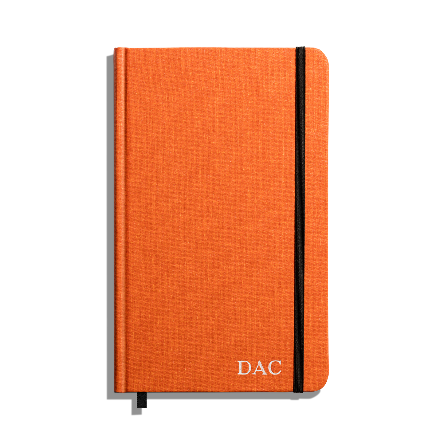 The Medium Hard Ruled Journal - Bold Orange by Shinola features a bold orange hardcover with “DAC” in white on the bottom right, a black elastic band, and ribbon bookmark. This American-made journal is set against a white background.