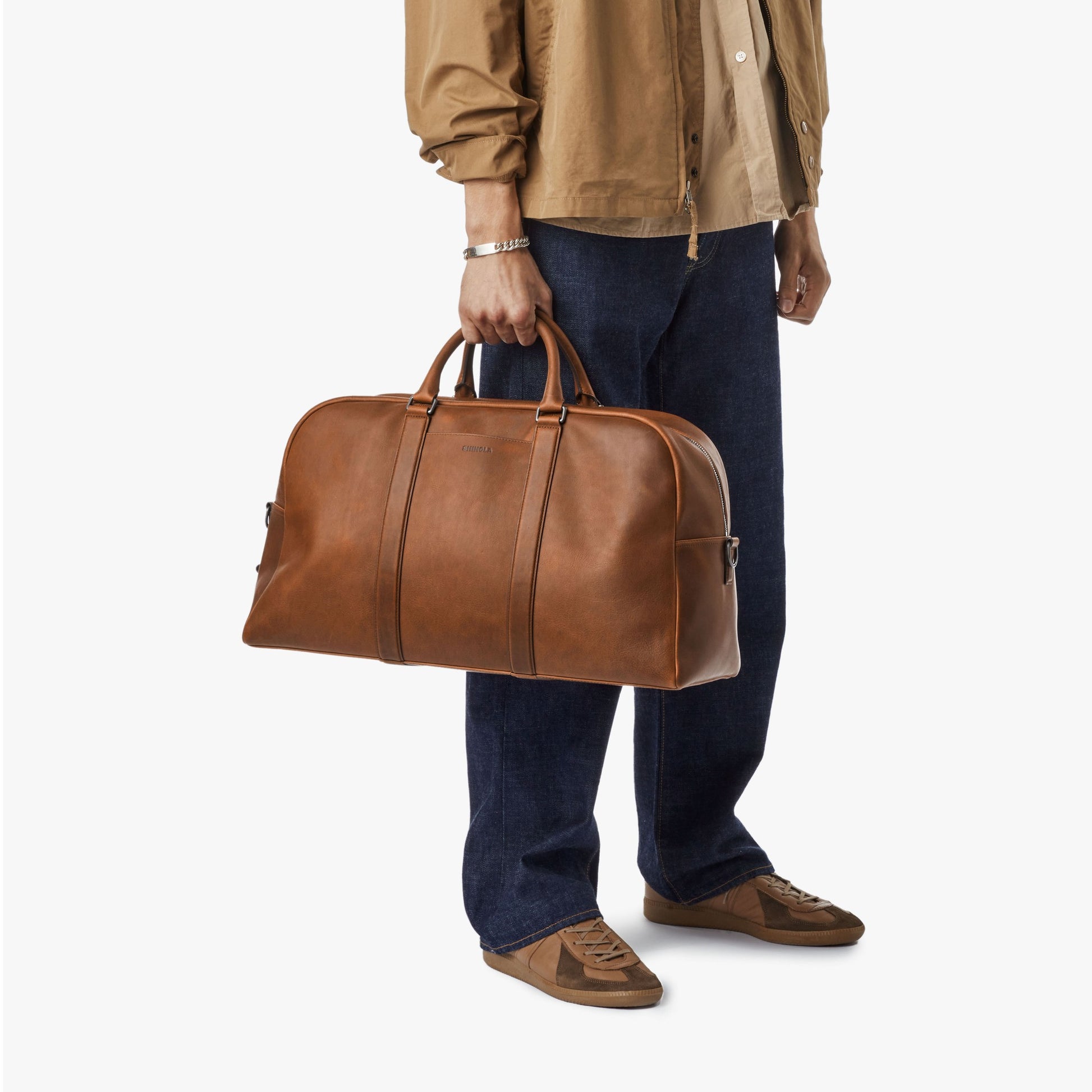 A person in a brown shirt, dark jeans, and brown shoes holds the Shinola Runwell Duffle Bag - Medium Brown by its handles against a plain white background.