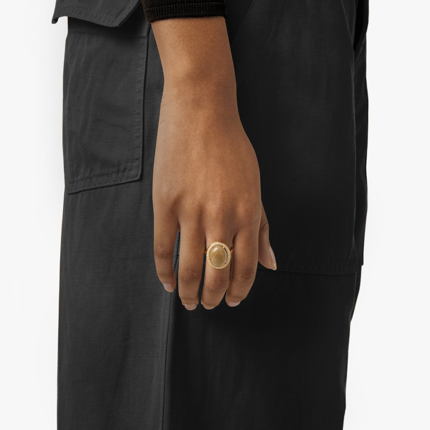 A close-up shows a hand wearing the Shinola Diamond Pave Oval Petoskey Ring in 14k yellow gold, resting against a dark outfit. The focus is on the elegant ring and hand, with the background softly blurred.