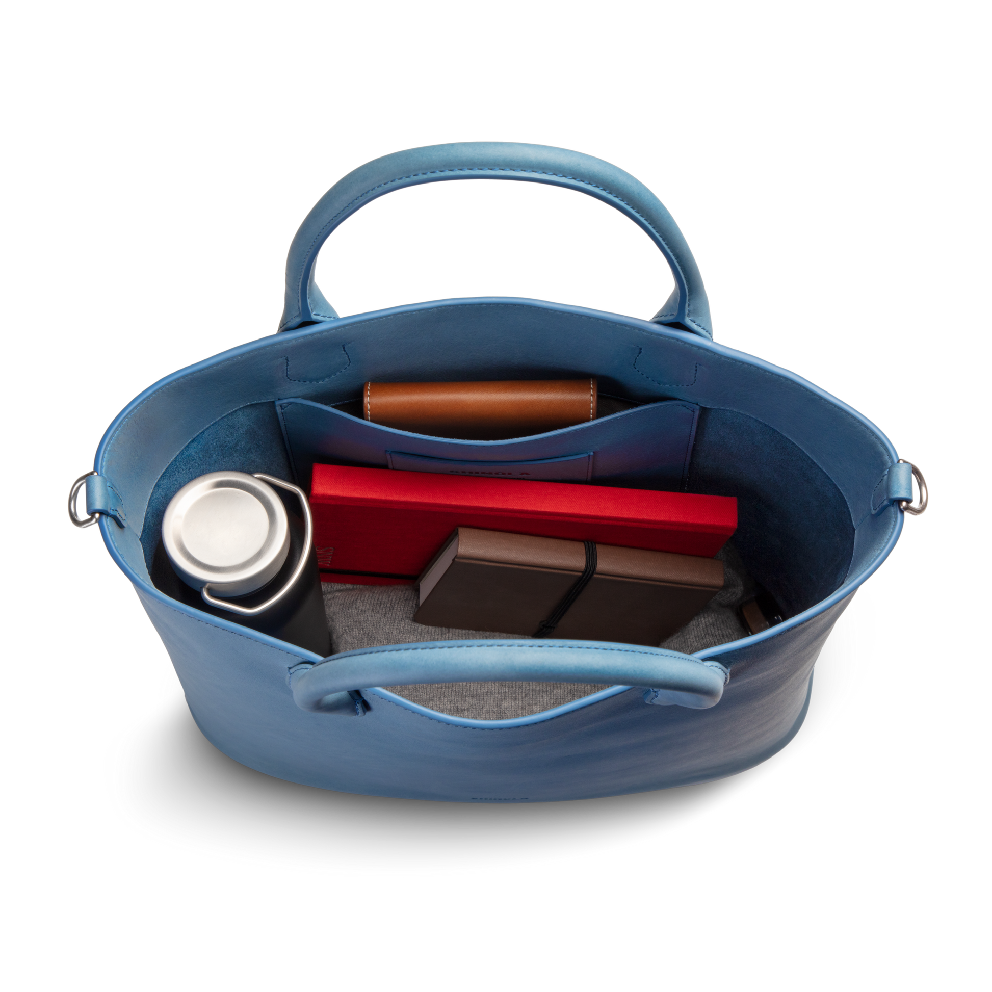 Top view of the Shinola 12 Hour Tote Bag - River in natural leather, open to show a stainless steel travel mug, a brown wallet, a red box, two smaller boxes, and a gray fabric item inside.