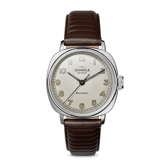 The Shinola Mechanic Watch - Alabaster features a hand-wound movement, round white dial with gold numerals, silver case, and brown leather strap. “MECHANIC” is above 6 o’clock. Displayed on a white background.