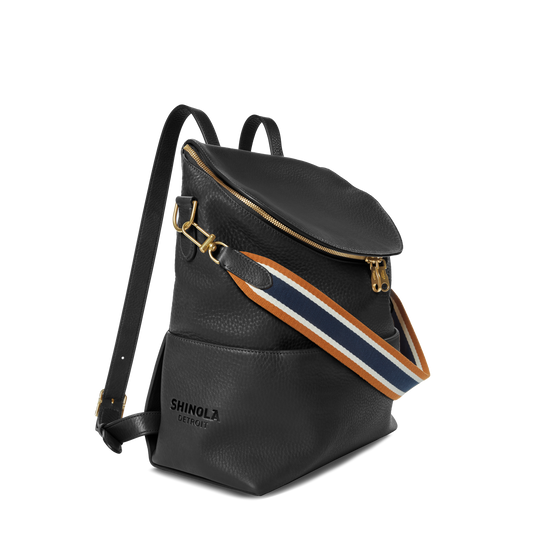 The Shinola Convertible Pocket Backpack - Brand Stripe / Black features gold hardware, a zippered top, and an adjustable navy, white, and brown striped strap. The front displays the Shinola Detroit logo—ideal for hands-free style.