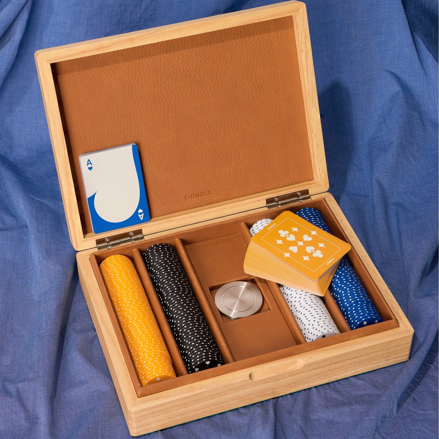 The Shinola Lacquered Wood Poker Set – Cream / Blue features a leather-lined box on blue fabric, with compartments for custom yellow, black, and white chips, a dealer button, yellow cards, and a single blue ace.