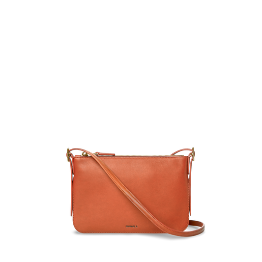 The Small Bixby Crossbody in Rust by Shinola is a small, rectangular bag crafted from natural leather with an adjustable strap and zippered top. The SHINOLA logo is printed near the bottom, making it a stylish everyday accessory.