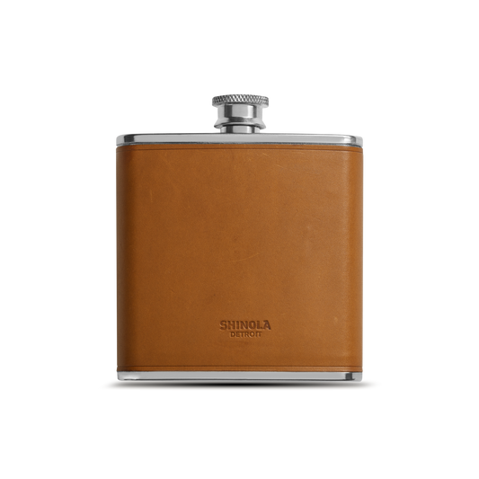 The Leather Wrapped Flask - Tan by Shinola features a stainless steel body with a tan Italian Vachetta leather wrap and screw cap, the Shinola name embossed on the front, displayed against a plain white background.