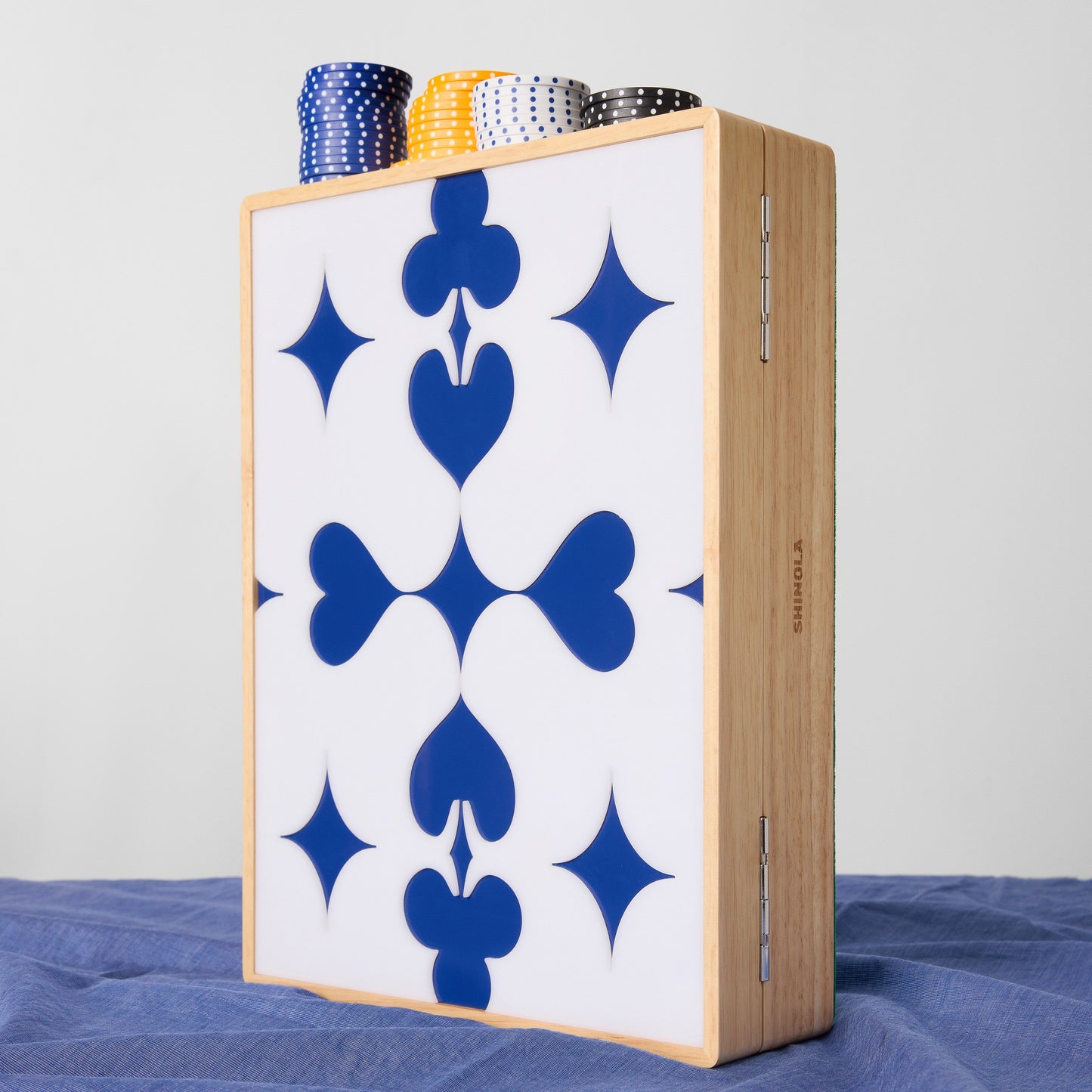 The Shinola Lacquered Wood Poker Set in Cream/Blue features a white panel with blue club and diamond shapes, an upright open leather-lined case, and stacks of custom poker chips displayed on a blue cloth.