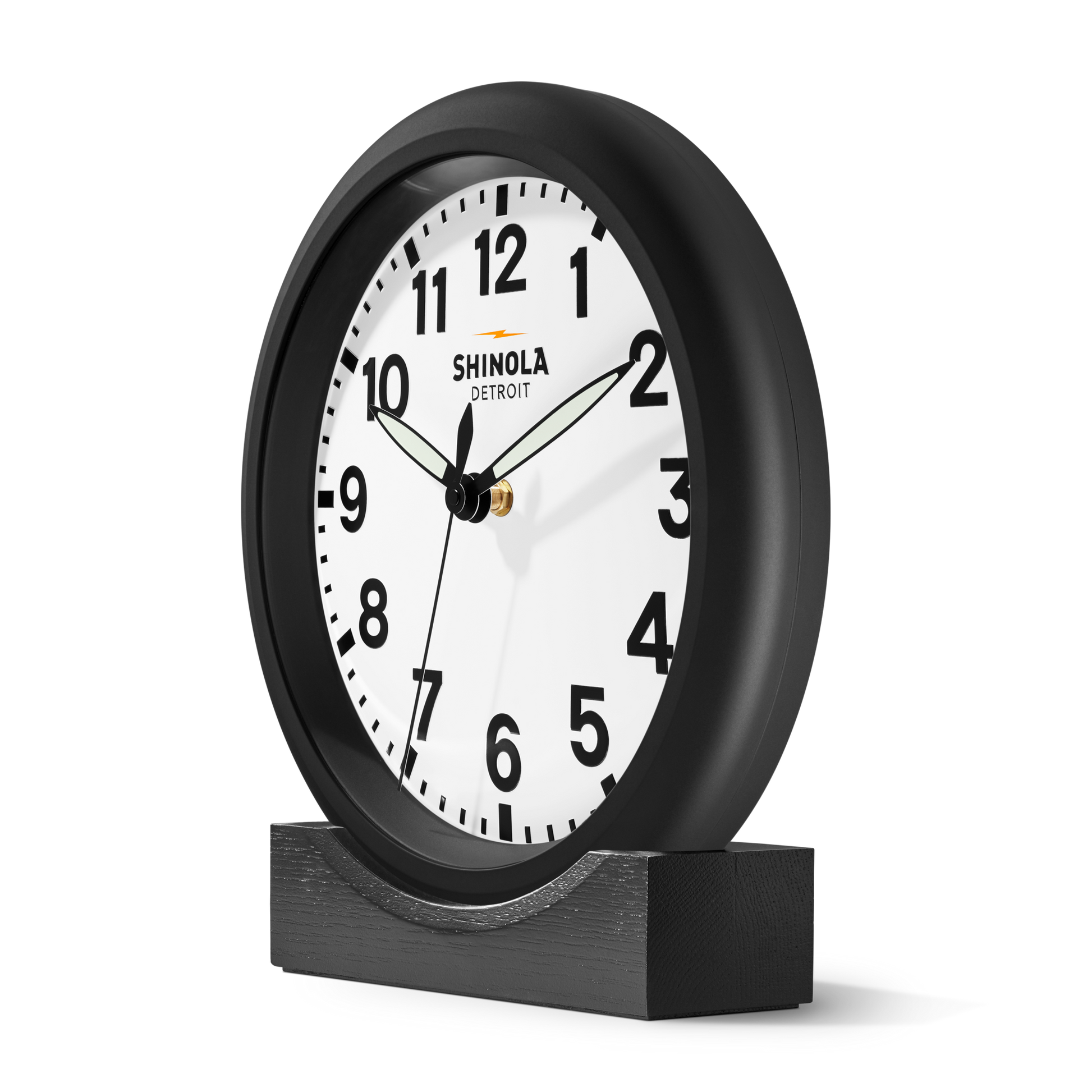 A Shinola 14" Runwell Clock in black with a white face, black numbers, and hands sits on a wooden stand. The clock face features Shinola branding and displays the time as 8:18:36.