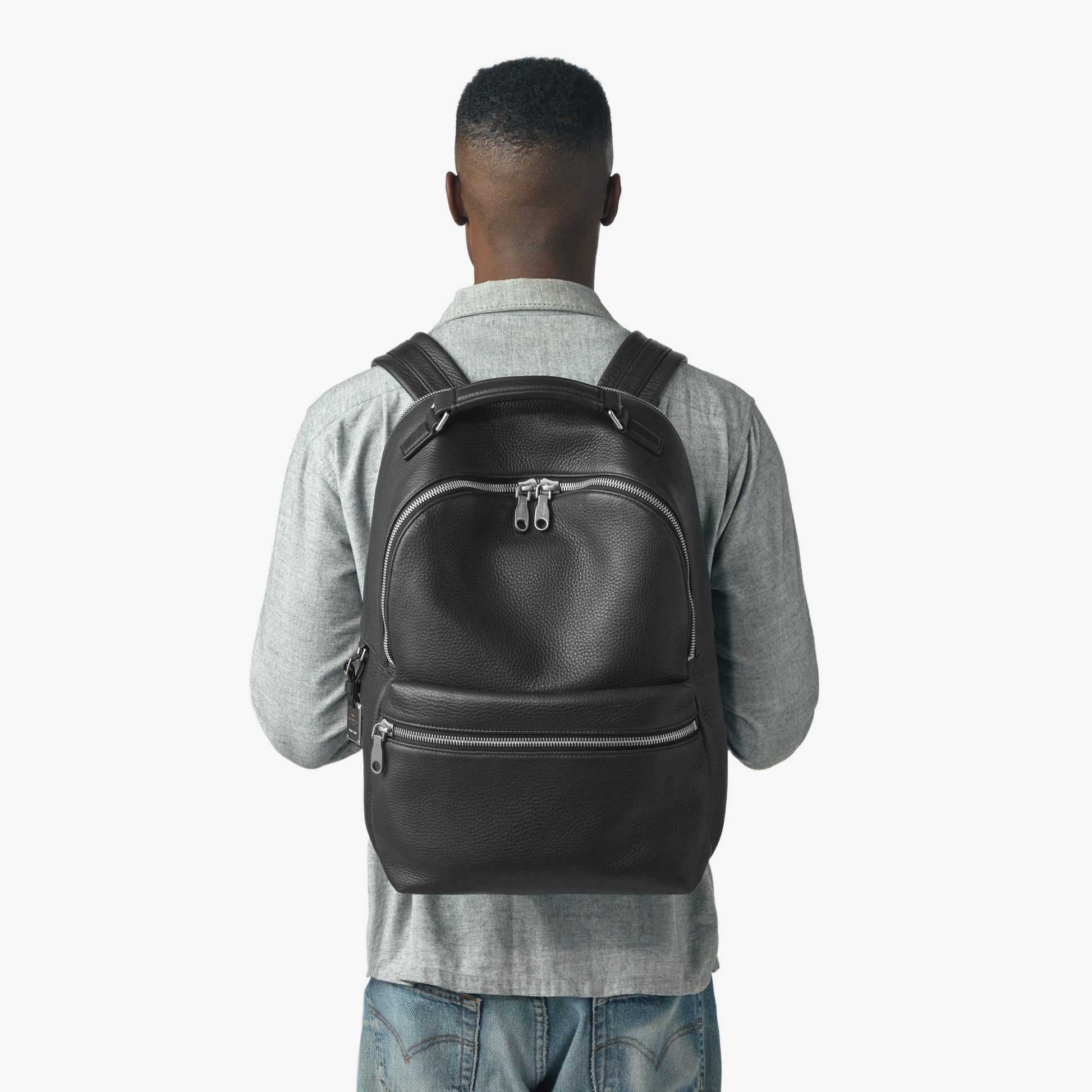 A person in a light gray shirt and blue jeans is seen from behind, carrying the Shinola Runwell Backpack - Black with multiple zippered compartments, set against a plain white background.