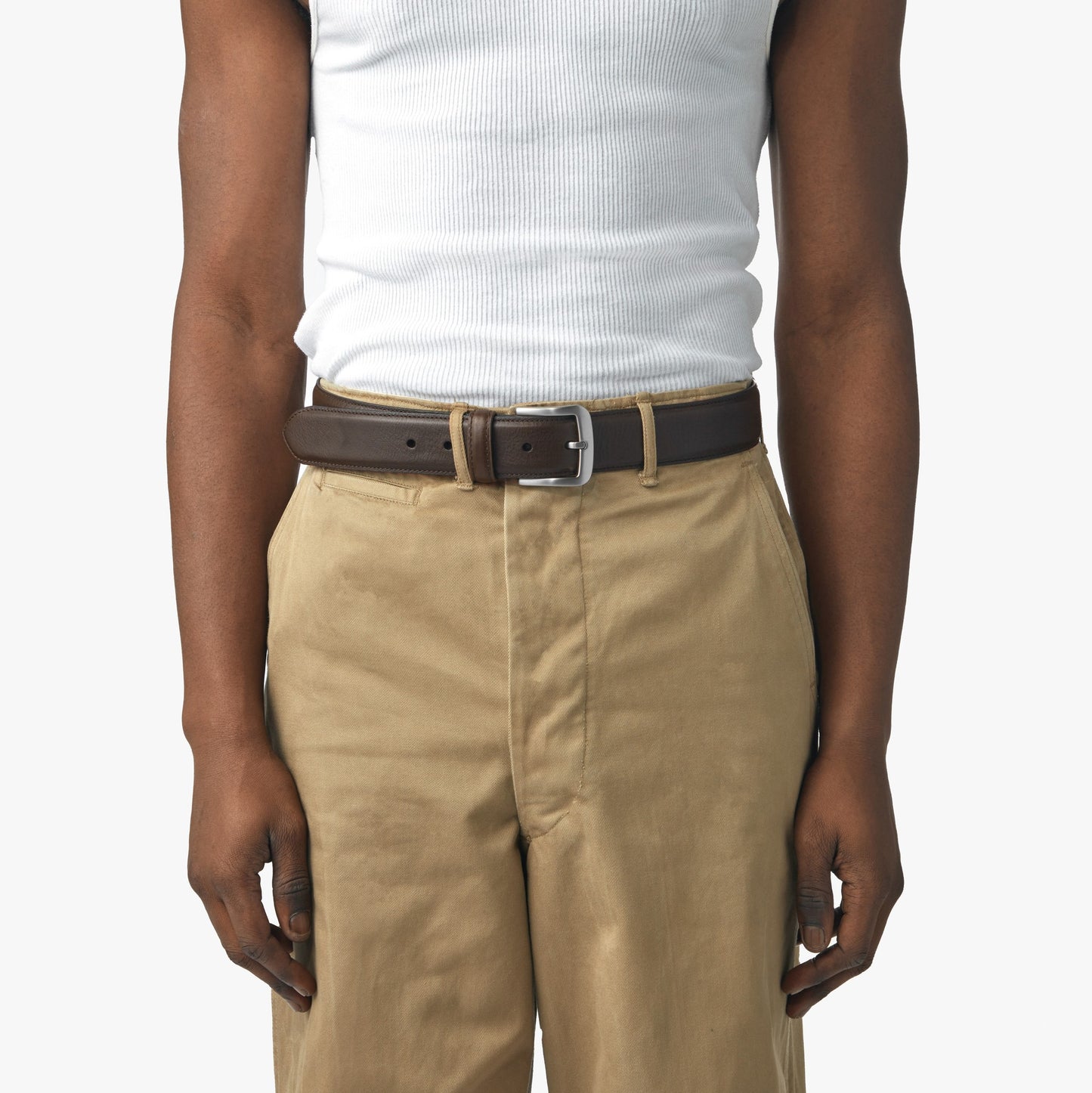 A person shown from shoulders to knees wears a white sleeveless ribbed shirt, tan pants, and the Shinola 1 1/2" Canfield Belt in Dark Brown against a plain background.