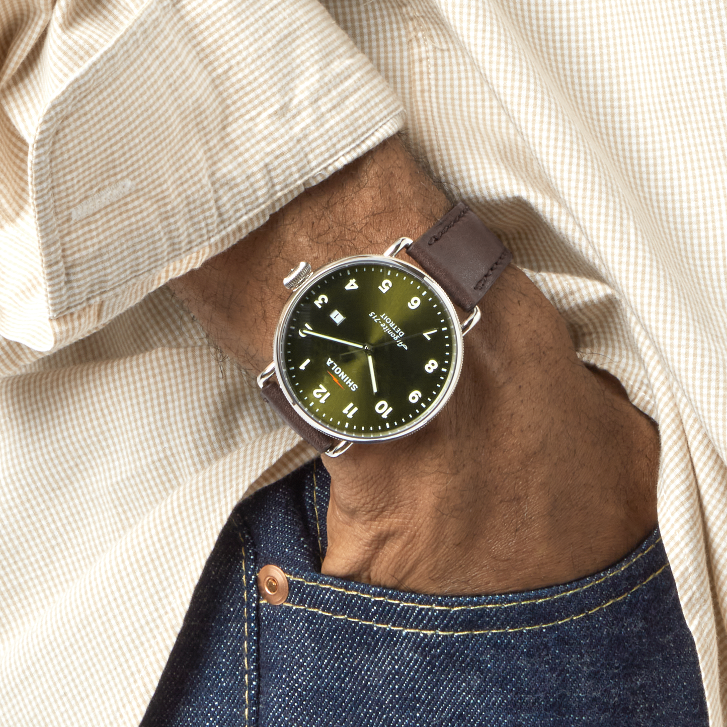 A person wearing a beige checkered shirt and blue jeans has their hand in their pocket, showing a Shinola Canfield Watch - Dark Olive with a brown leather strap, dark olive face with white numbers, and a sleek stainless steel case.