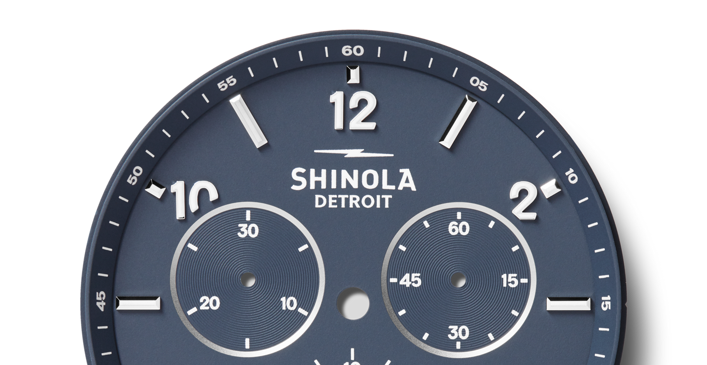 A close-up of the Shinola Canfield Sport Chronograph Watch in Midnight Blue features bold white hour markers, two subdials for seconds, and white minute markings along the edge. The watch hands are missing.