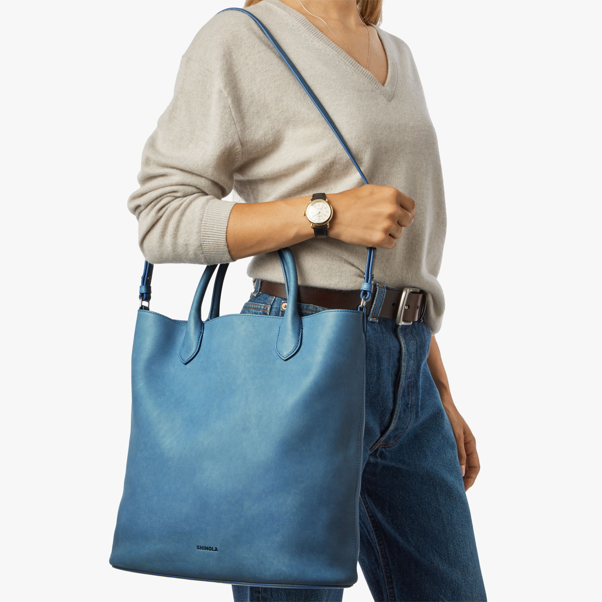 A woman in a light beige sweater and blue jeans carries the Shinola 12 Hour Tote Bag - River over her shoulder. She finishes her look with a wristwatch and brown belt, standing against a plain white background.