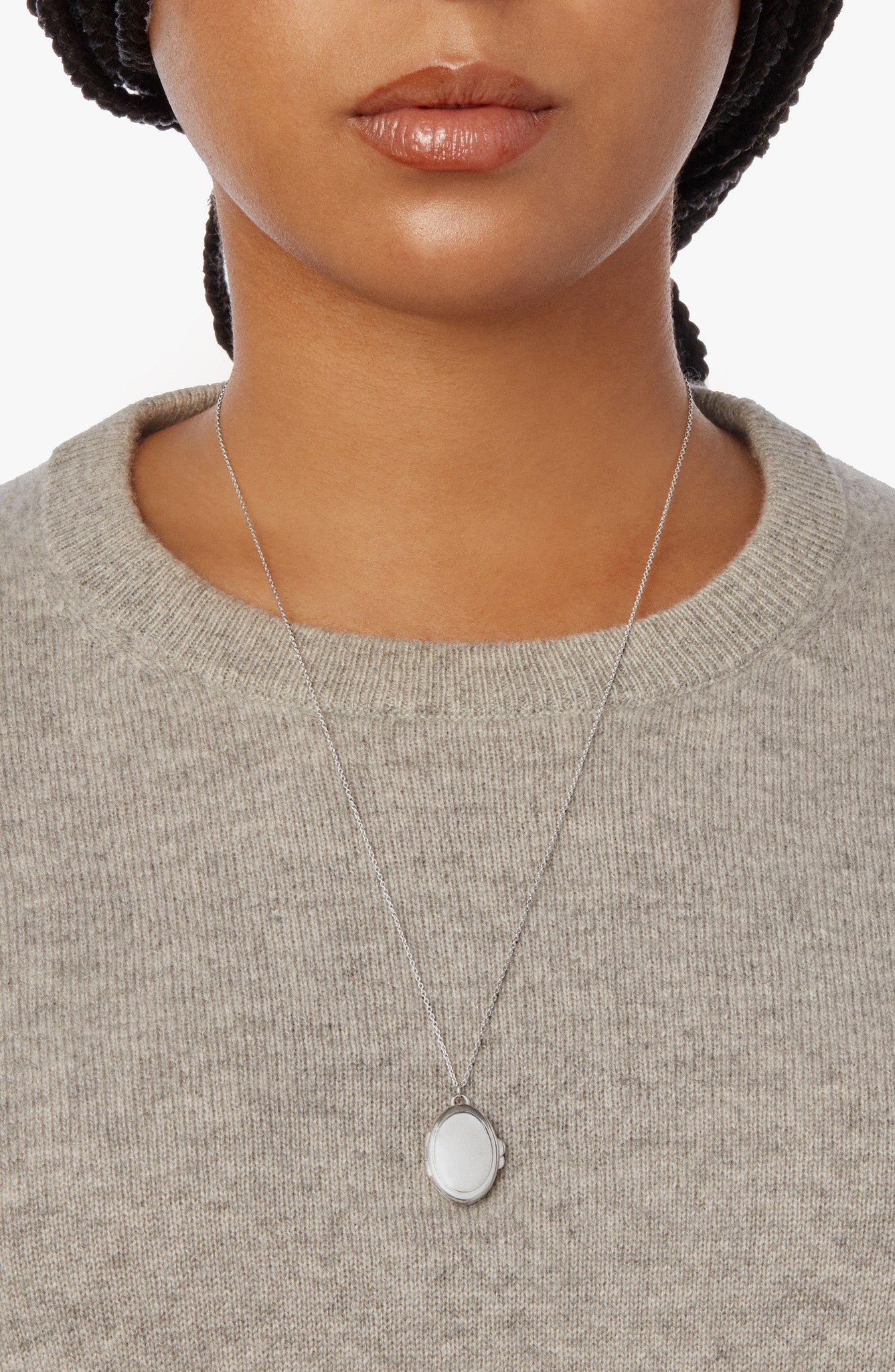 A person wears a light gray knit sweater and the Shinola Heirloom Statement Locket Necklace in sterling silver, featuring an oval white pendant, shown from shoulders to just above the lips.