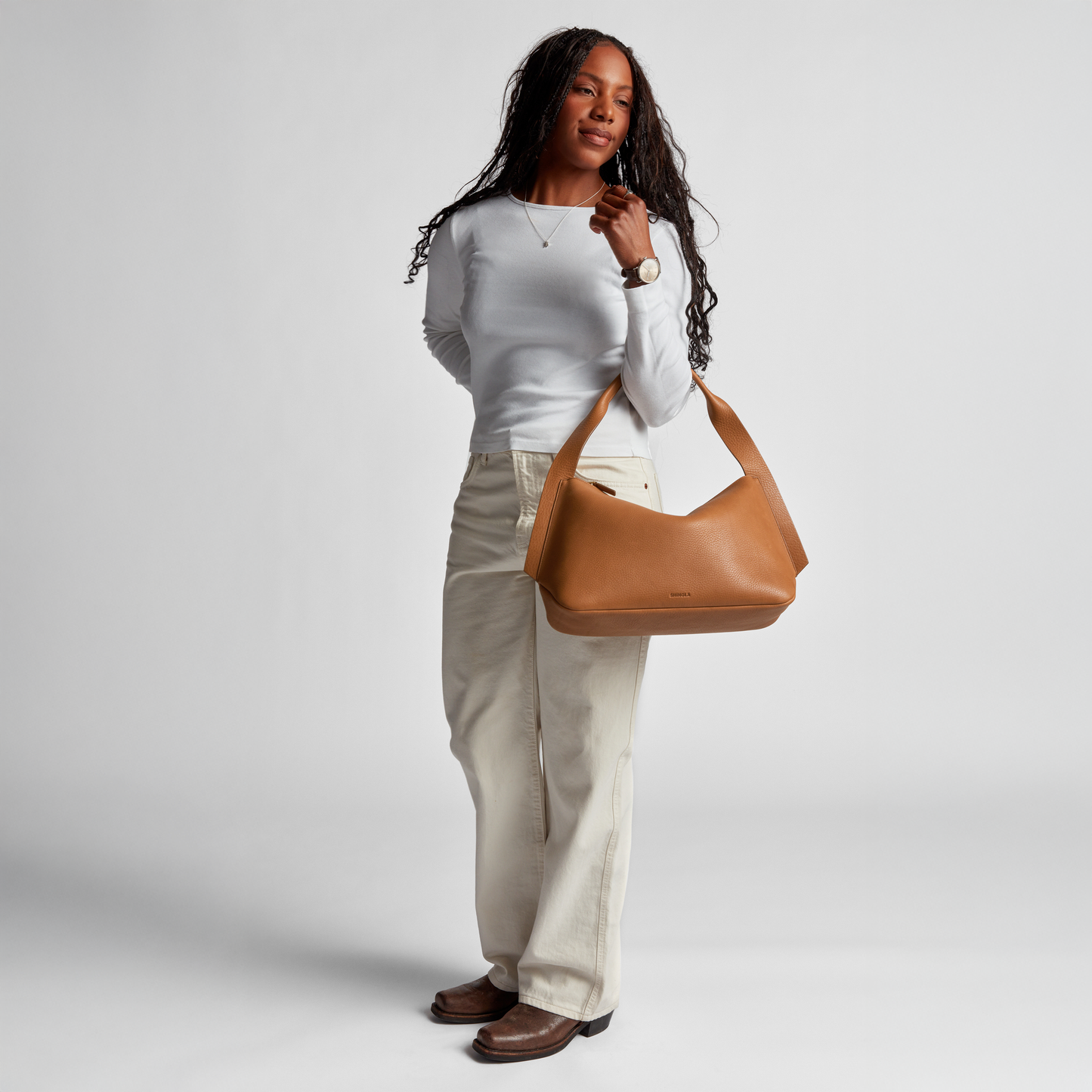 A woman with long curly hair smiles as she holds the Shinola Medium Derby Shoulder Bag - Tan, wearing a white long-sleeve shirt, cream-colored pants, and brown shoes against a plain light background.