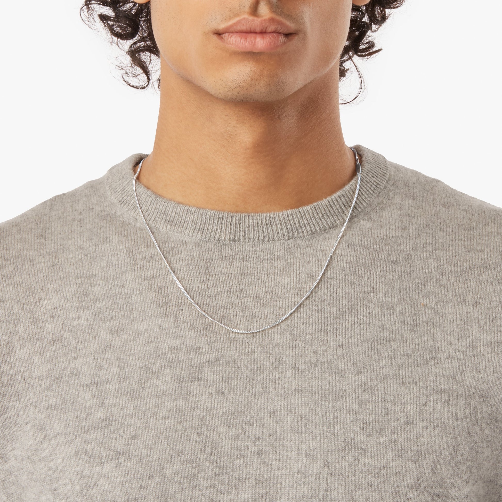 A person wearing a light gray knit sweater and the Shinola Simple Curb Chain Necklace in sterling silver, shown from shoulders to just above the lips against a plain white background.