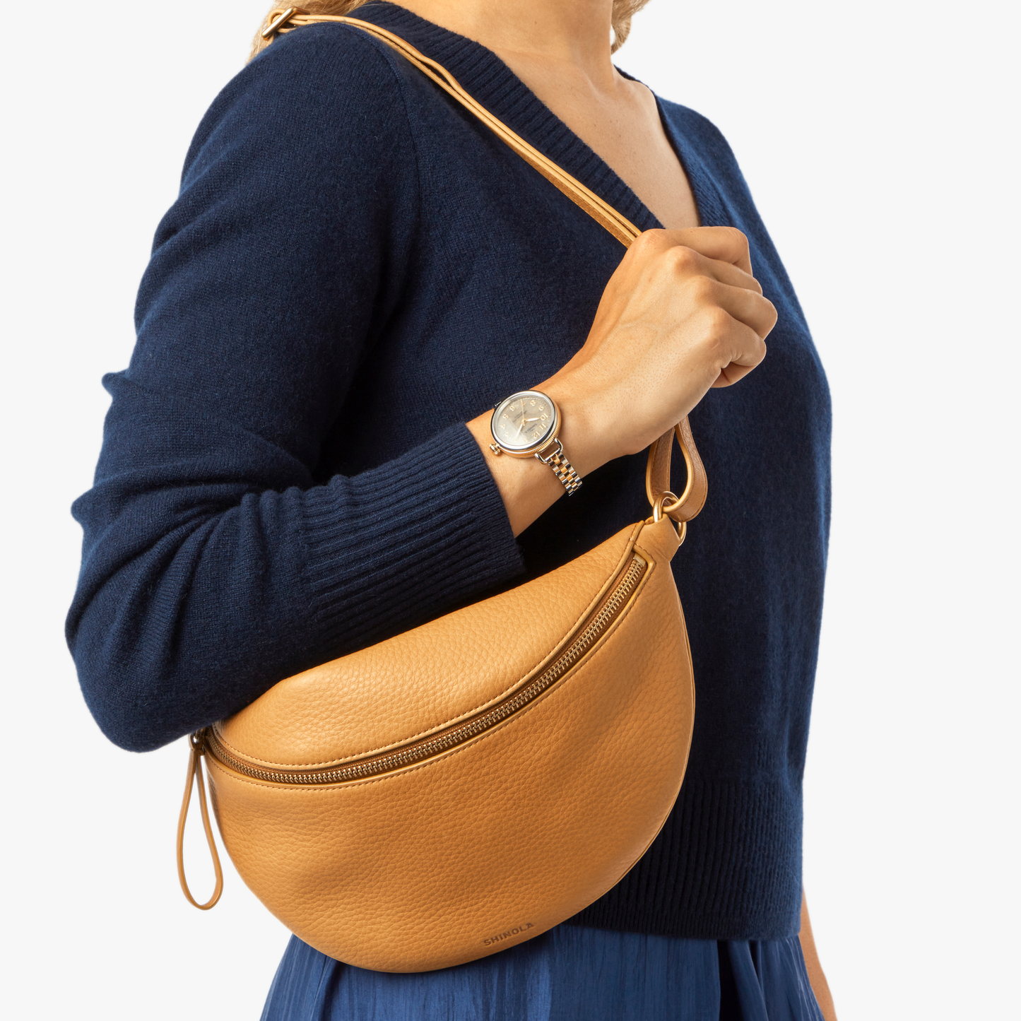 A person wearing a navy sweater and silver wristwatch holds the Shinola Sling Bag - Honey, featuring an adjustable leather strap and zipper, against a plain white background.