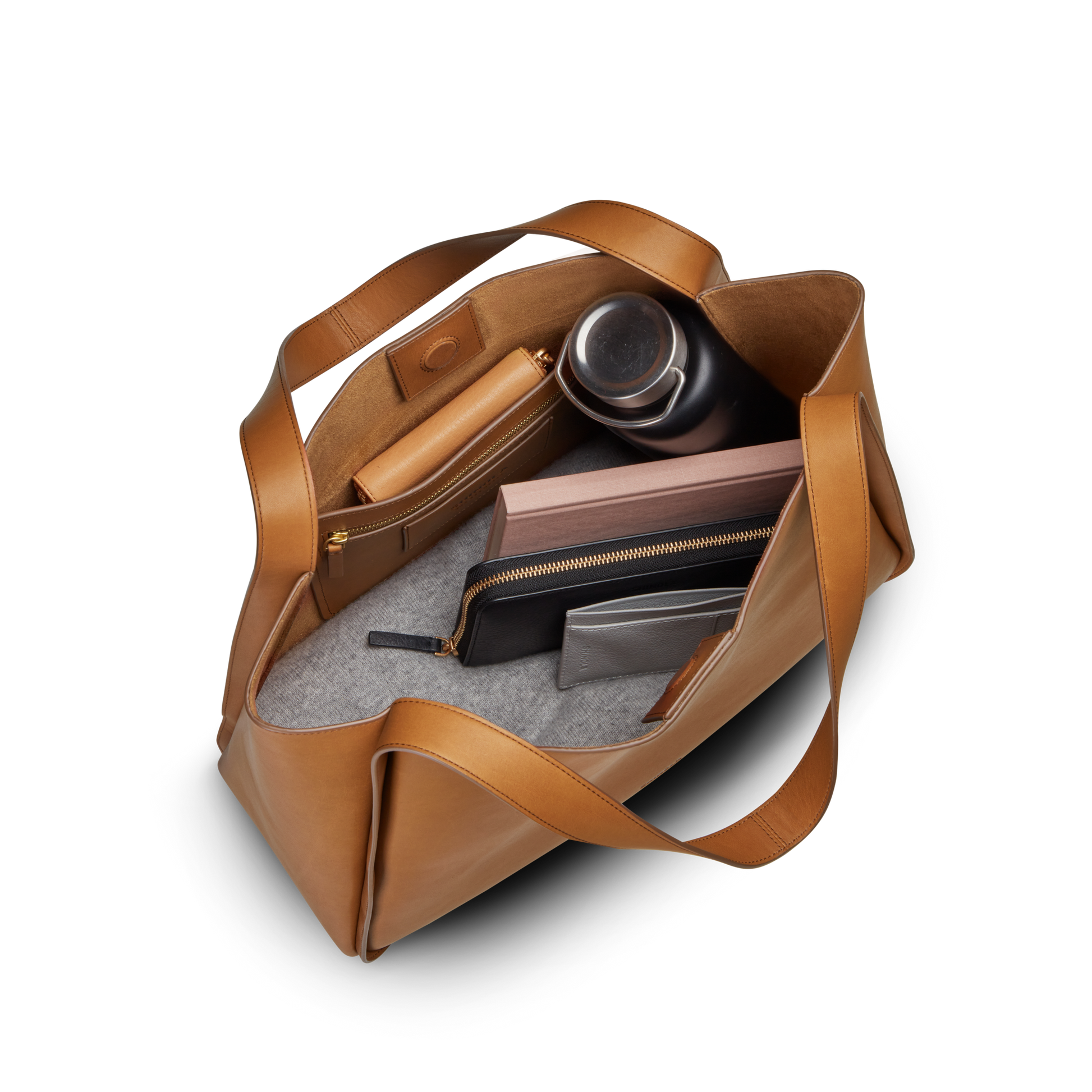 A top-down view of the Shinola Derby Tote Bag - Tan, crafted from natural leather with gray lining, neatly holding a black water bottle, several zippered pouches, a notebook, and a wallet.