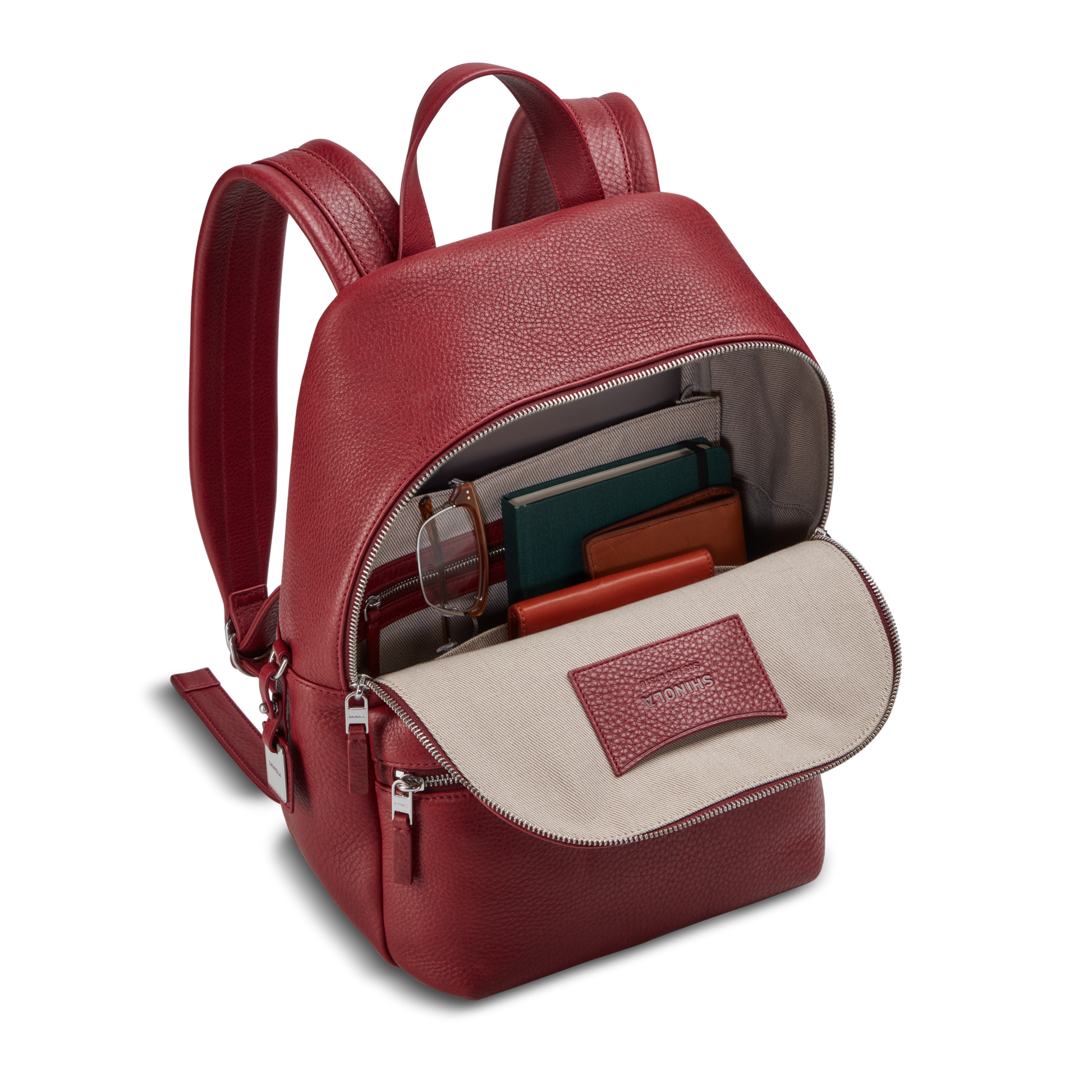 The Small Runwell Backpack - Ruby by Shinola, made from natural grain leather, sits unzipped with glasses, notebooks, and a wallet neatly arranged inside against a neutral background.