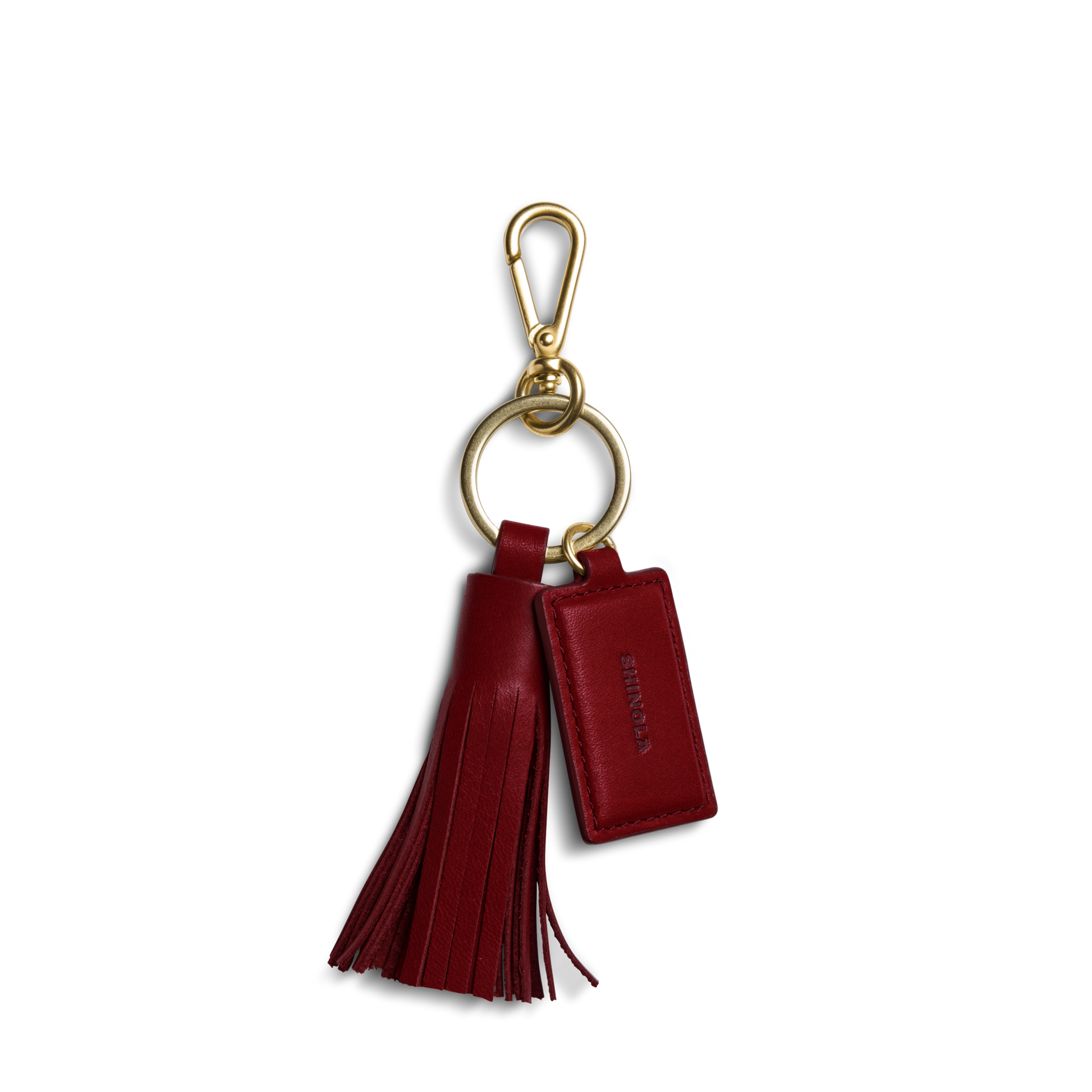 The Leather Tassel Keychain - Ruby by Shinola features a gold keyring with clasp, a red natural leather tassel, and a rectangular monogrammable tag embossed with SHINOLA. Shown on a plain white background.