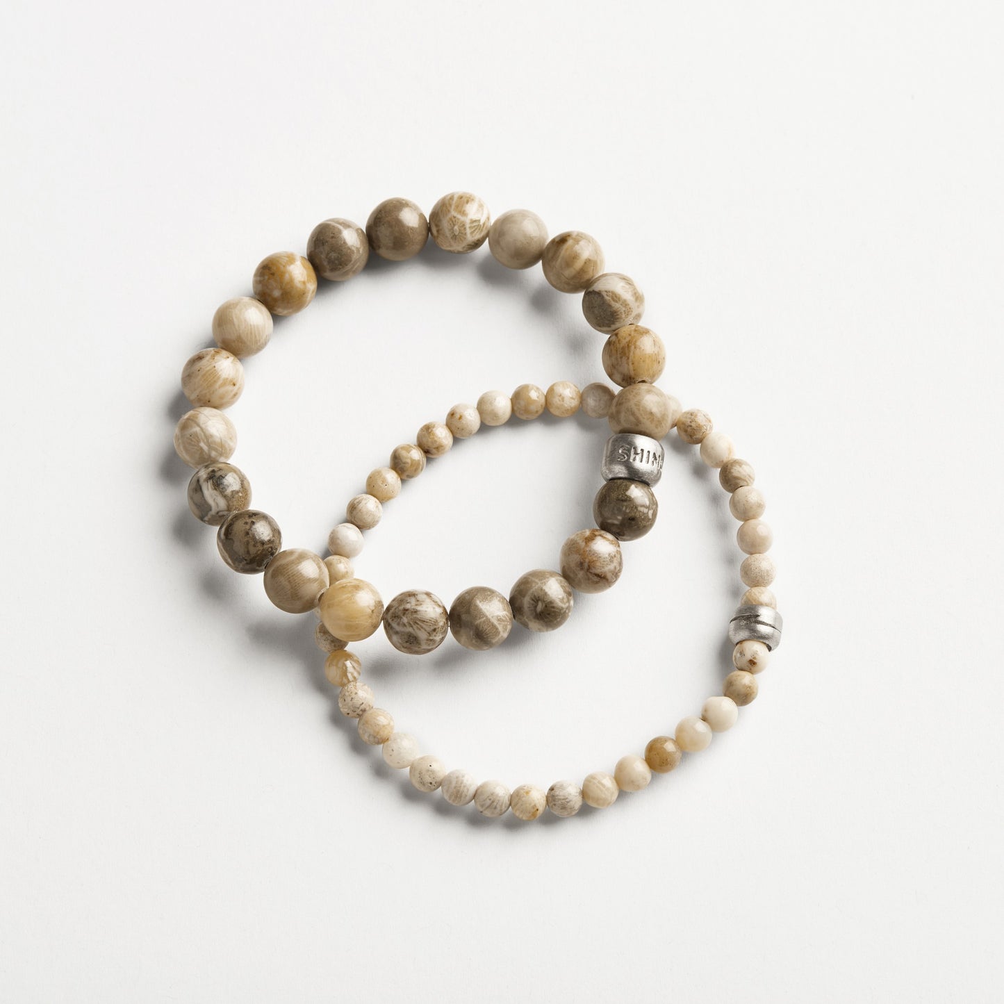 Two stretch bracelets rest on a white surface: an 8mm Beaded Petoskey Bracelet by Shinola with large, earth-toned beads, and another with smaller beads plus a sterling silver charm engraved with an inspiring word.