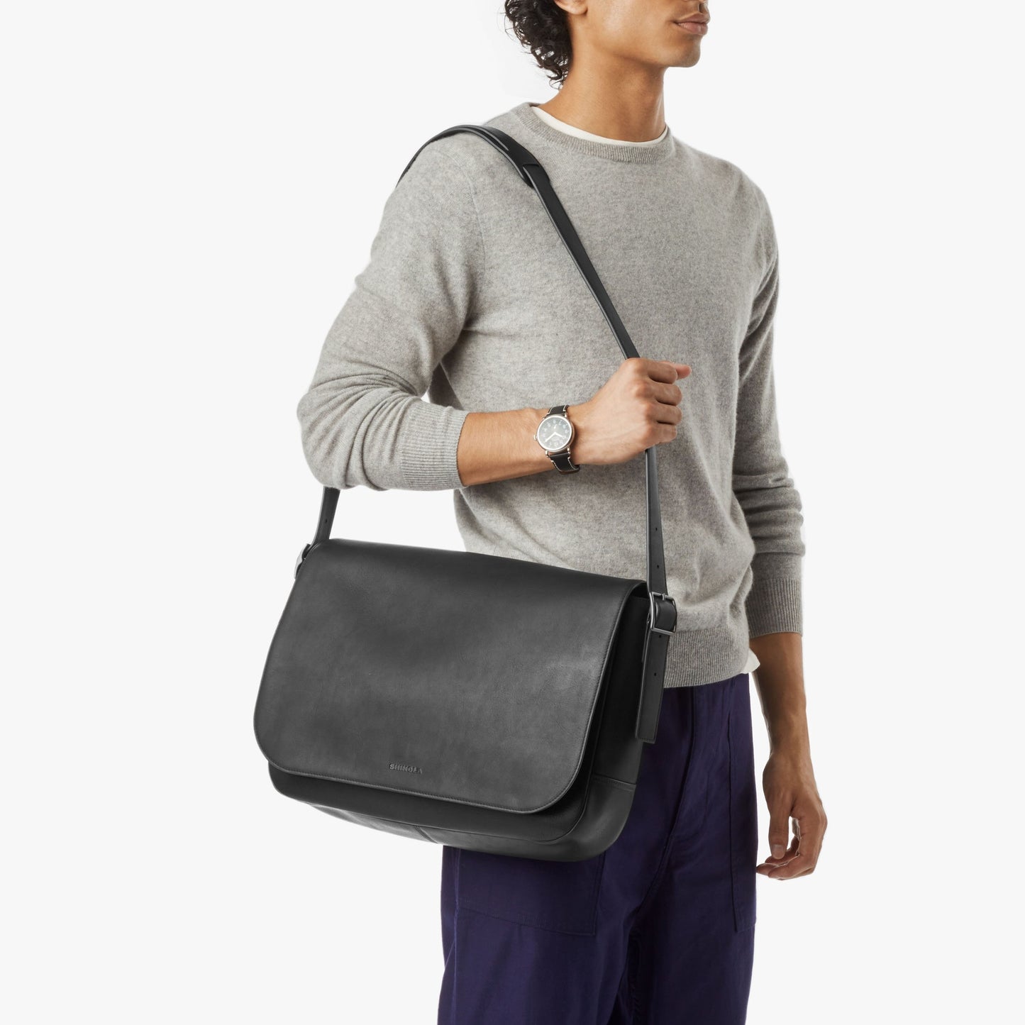 A person in a light gray sweater and dark blue pants carries the Shinola Runwell Messenger Bag - Black, featuring an adjustable crossbody strap, flap closure, and padded laptop sleeve. They also wear a wristwatch.
