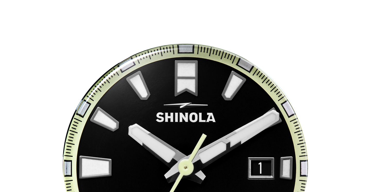 Close-up of the top half of a black Shinola Forged Carbon Monster Watch face with bold white hour markers, logo, and date window at 3 o’clock. Minute and hour hands show 4:02. Features a durable forged carbon case.