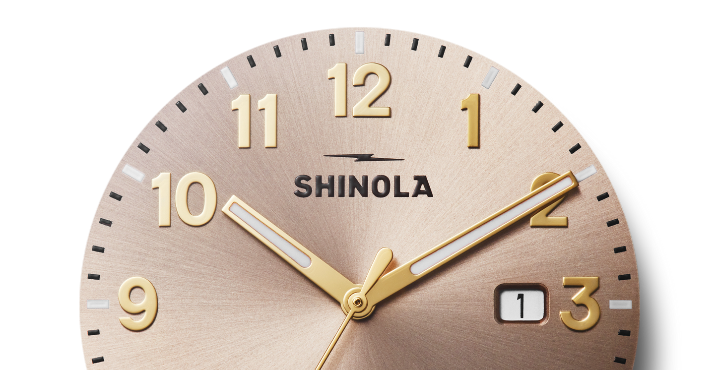 Close-up of the Shinola Detrola Watch - Nude, featuring gold numbers and hands, a small date window near 3, and a brushed metallic face—highlighting classic American design. Time displayed is about 10:10.