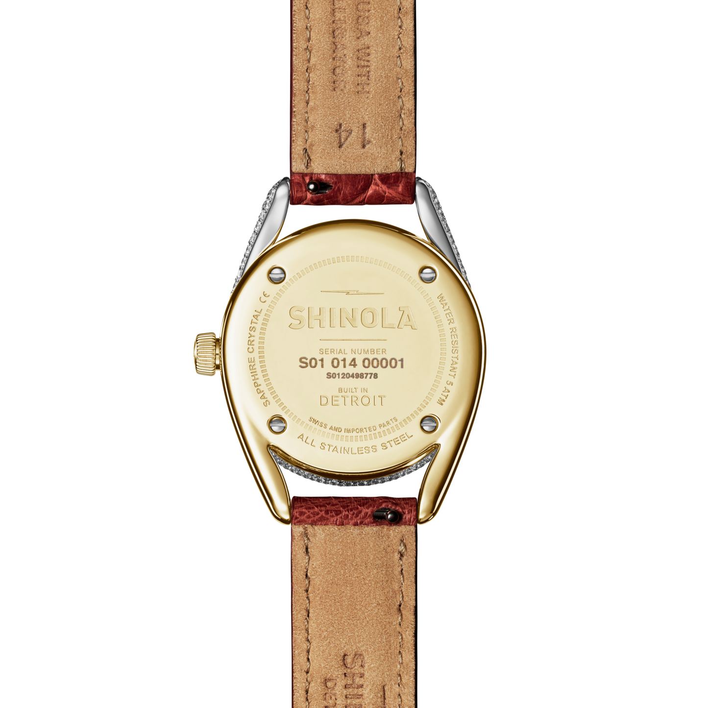 A close-up of the back of the Shinola Diamond Derby Watch shows a gold-toned, two-tone horseshoe-shaped case engraved with the brand name and “Built in Detroit.” The auburn alligator strap is also visible.