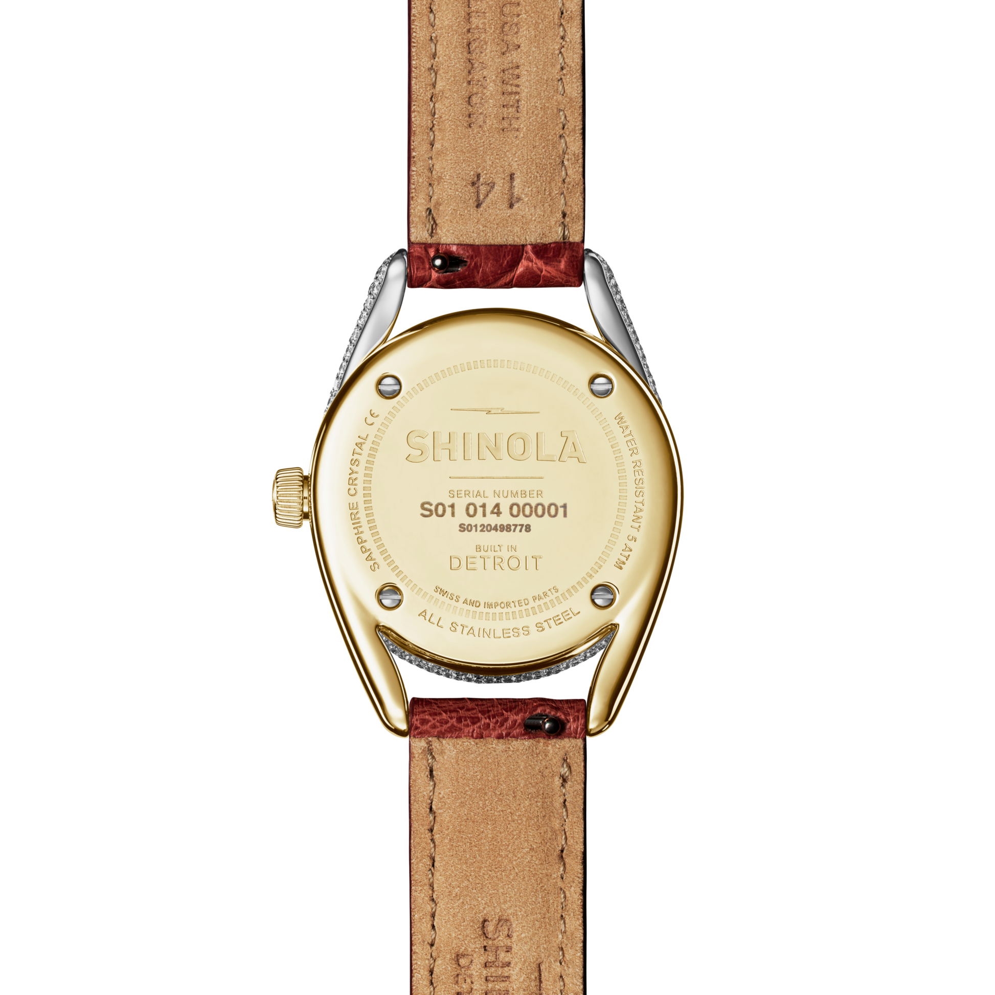 A close-up of the back of the Shinola Diamond Derby Watch shows a gold-toned, two-tone horseshoe-shaped case engraved with the brand name and “Built in Detroit.” The auburn alligator strap is also visible.