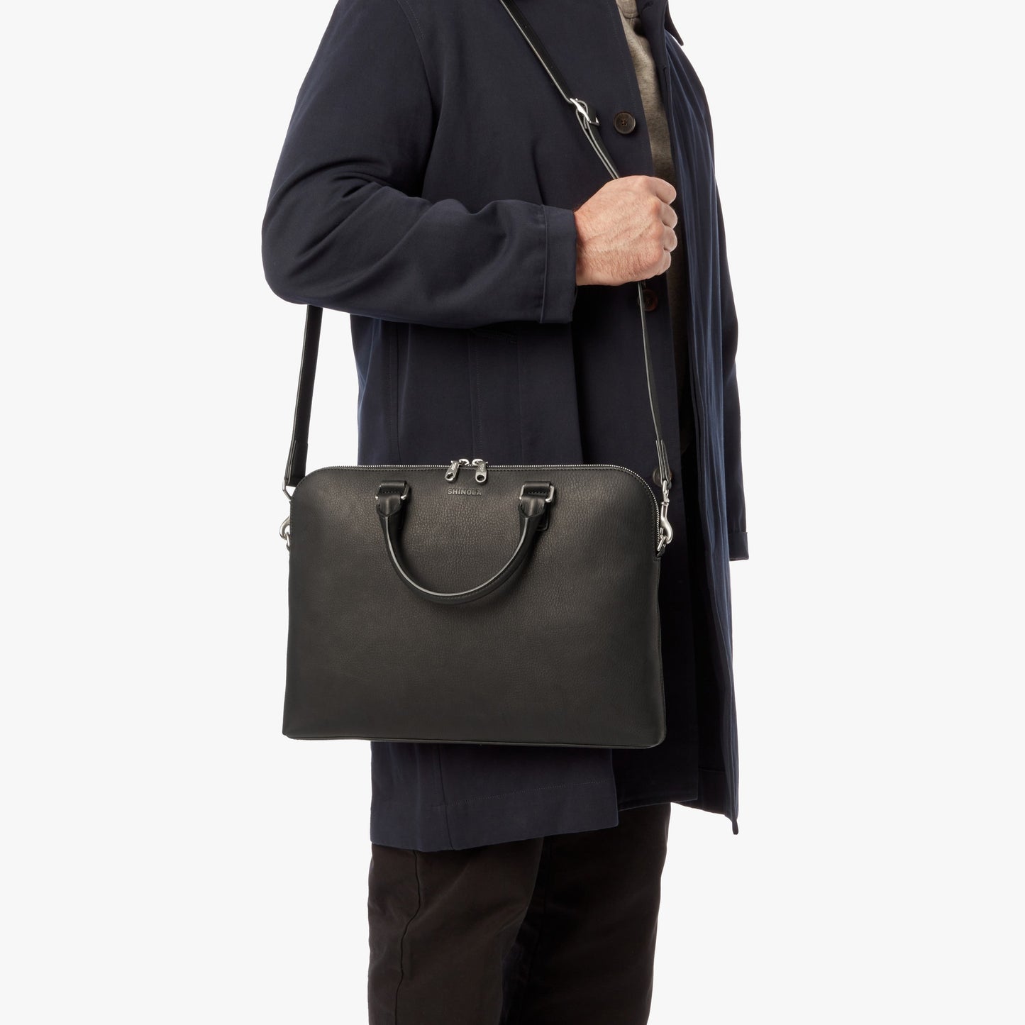 A person in a dark coat holds the Shinola Portfolio Briefcase in black European grain leather, featuring a zip closure and long strap, displayed against a plain white background.